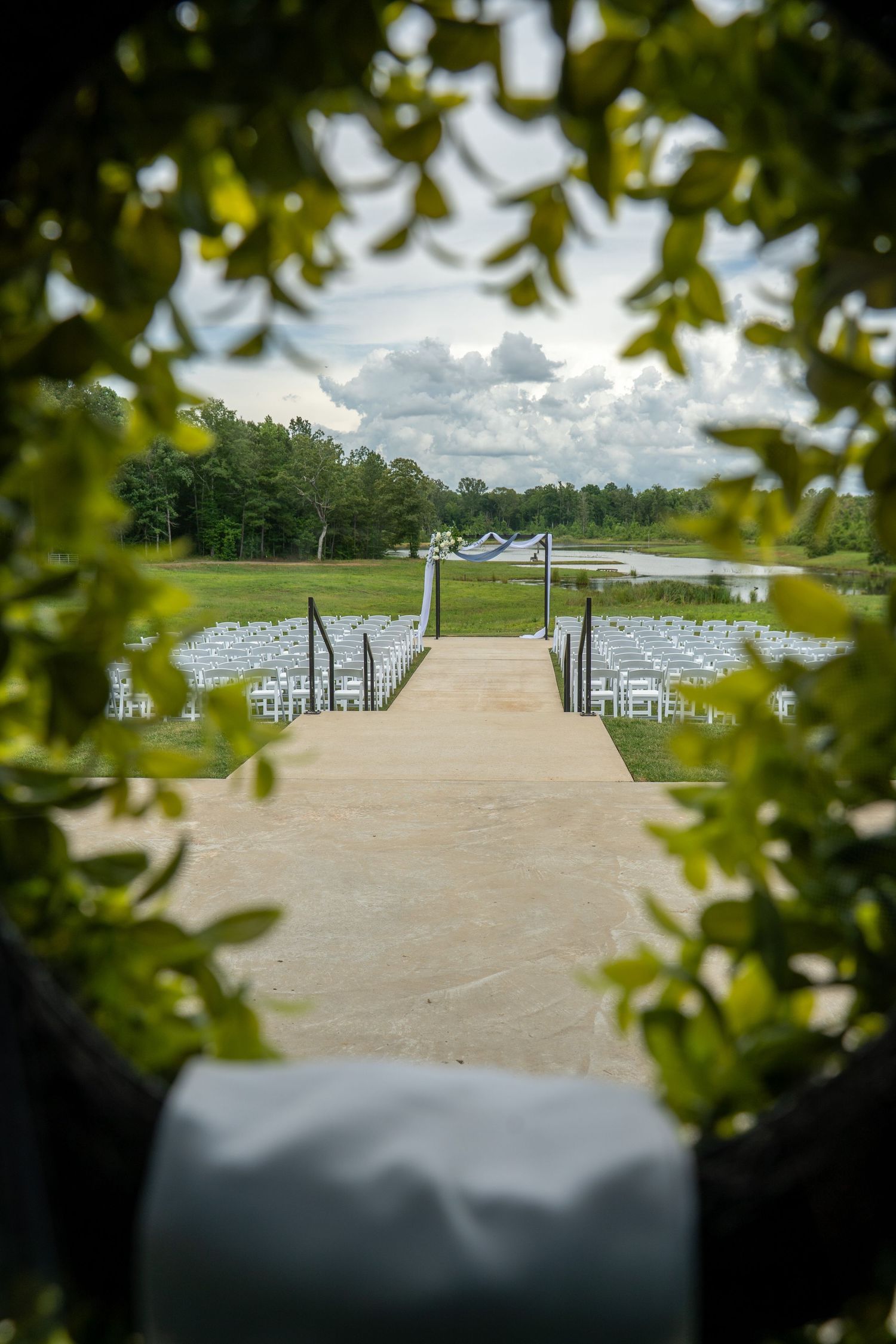 Outdoor wedding ceremony setup with white chairs along a path, framed by lush green foliage and a scenic pond backdrop.