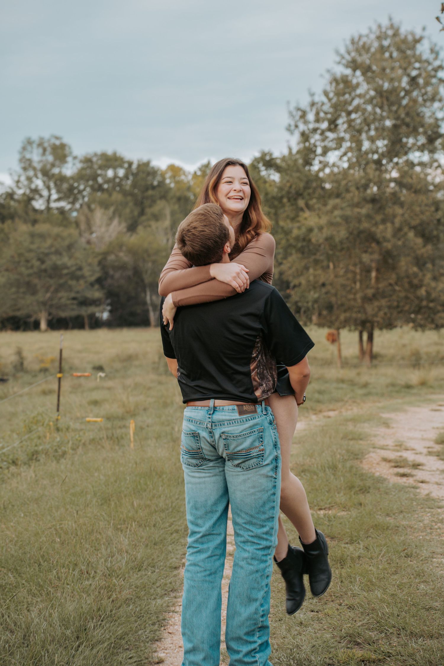 A joyful woman laughs while being lifted in a warm embrace outdoors on a rural dirt path surrounded by green trees.