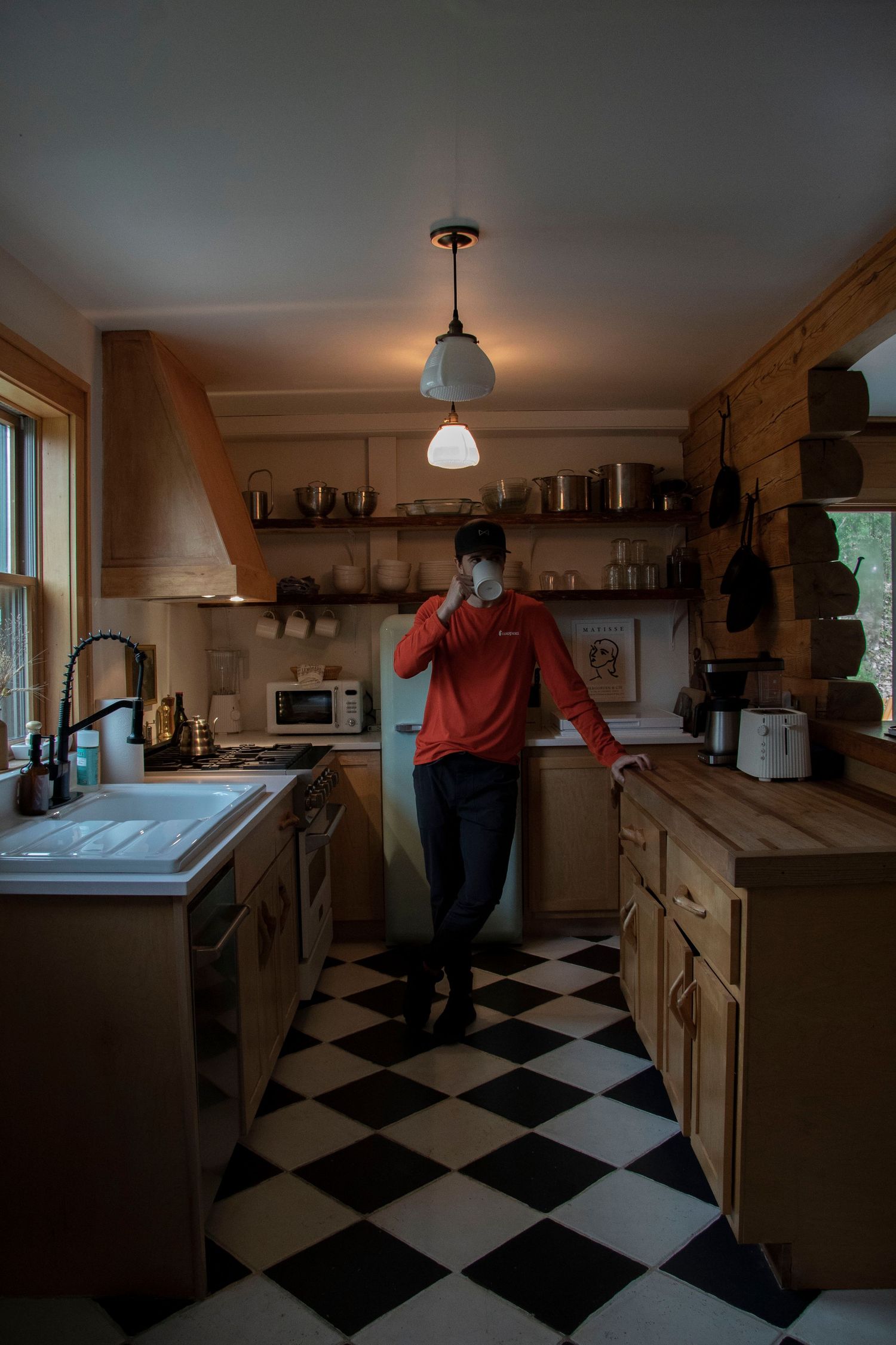 Man in red hoodie standing in a rustic kitchen with checkered floors, wooden cabinets, and warm pendant lighting.