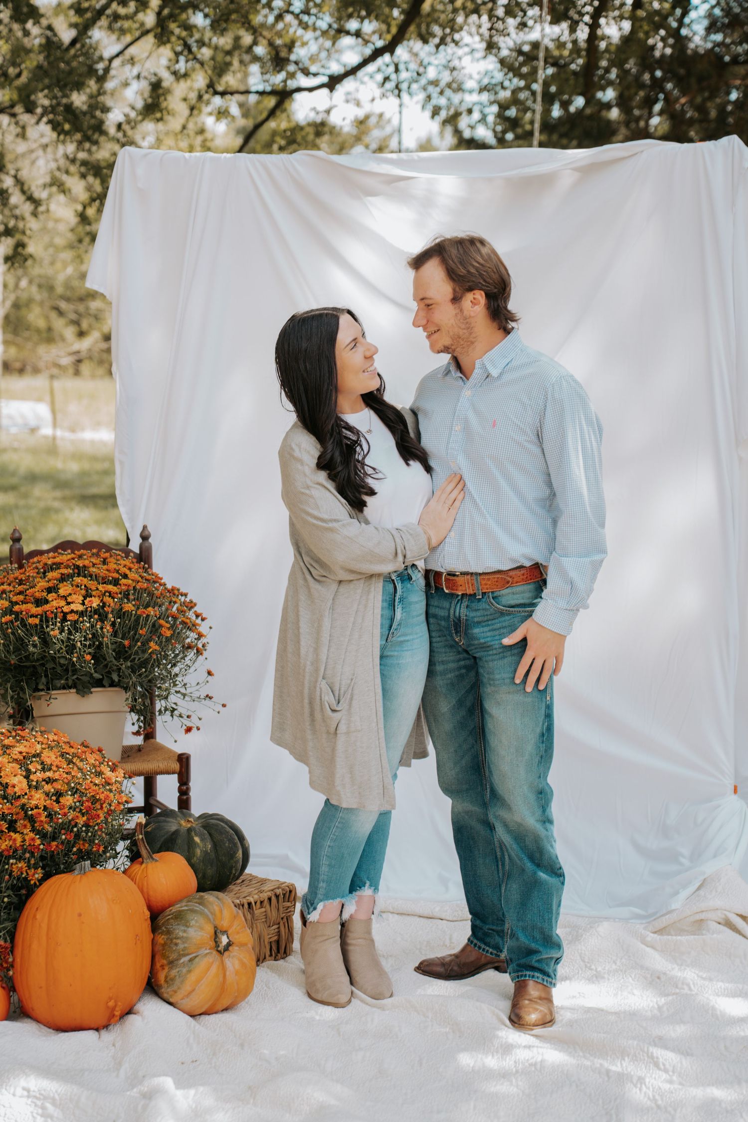 Couple sharing a romantic moment in front of white backdrop with fall pumpkins and orange mums in outdoor autumn setting.