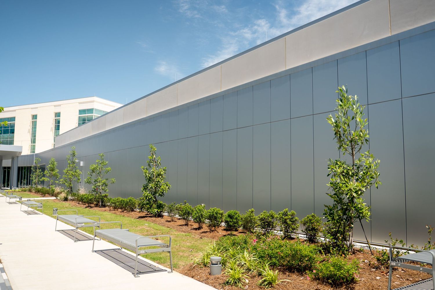 Modern commercial building exterior with gray panel facade, landscaped courtyard featuring young trees and shrubs under blue sky.