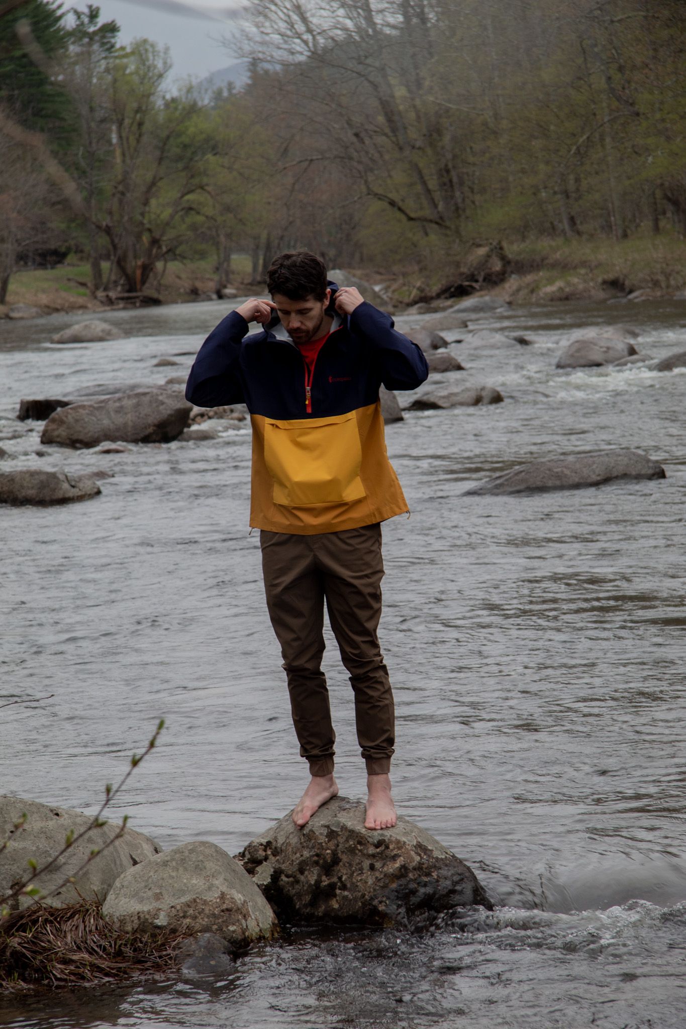 Barefoot man standing on river rocks wearing yellow and navy pullover jacket with brown jogger pants outdoors.