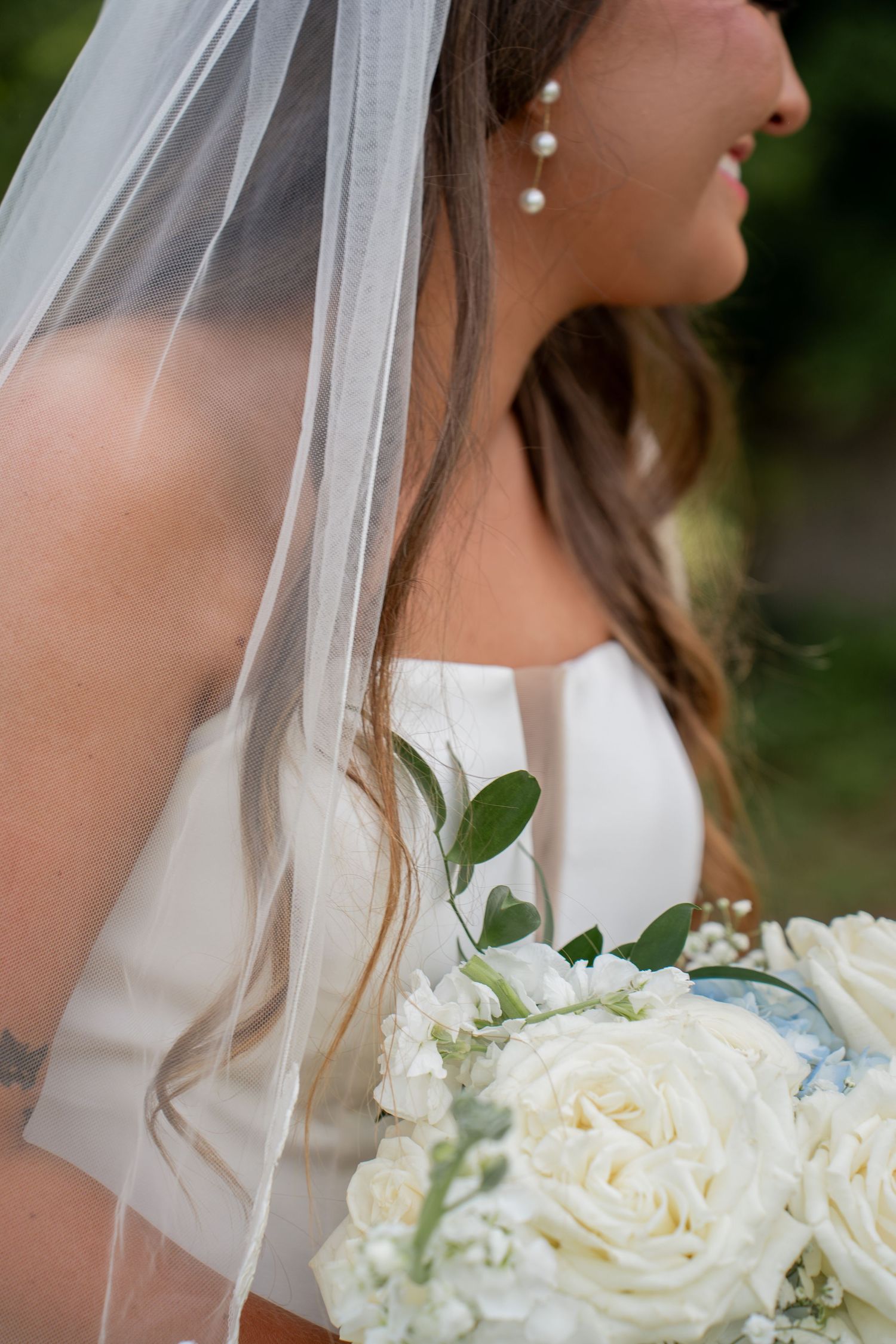 Bride in white strapless gown with veil and pearl earrings holding white rose bouquet with greenery.