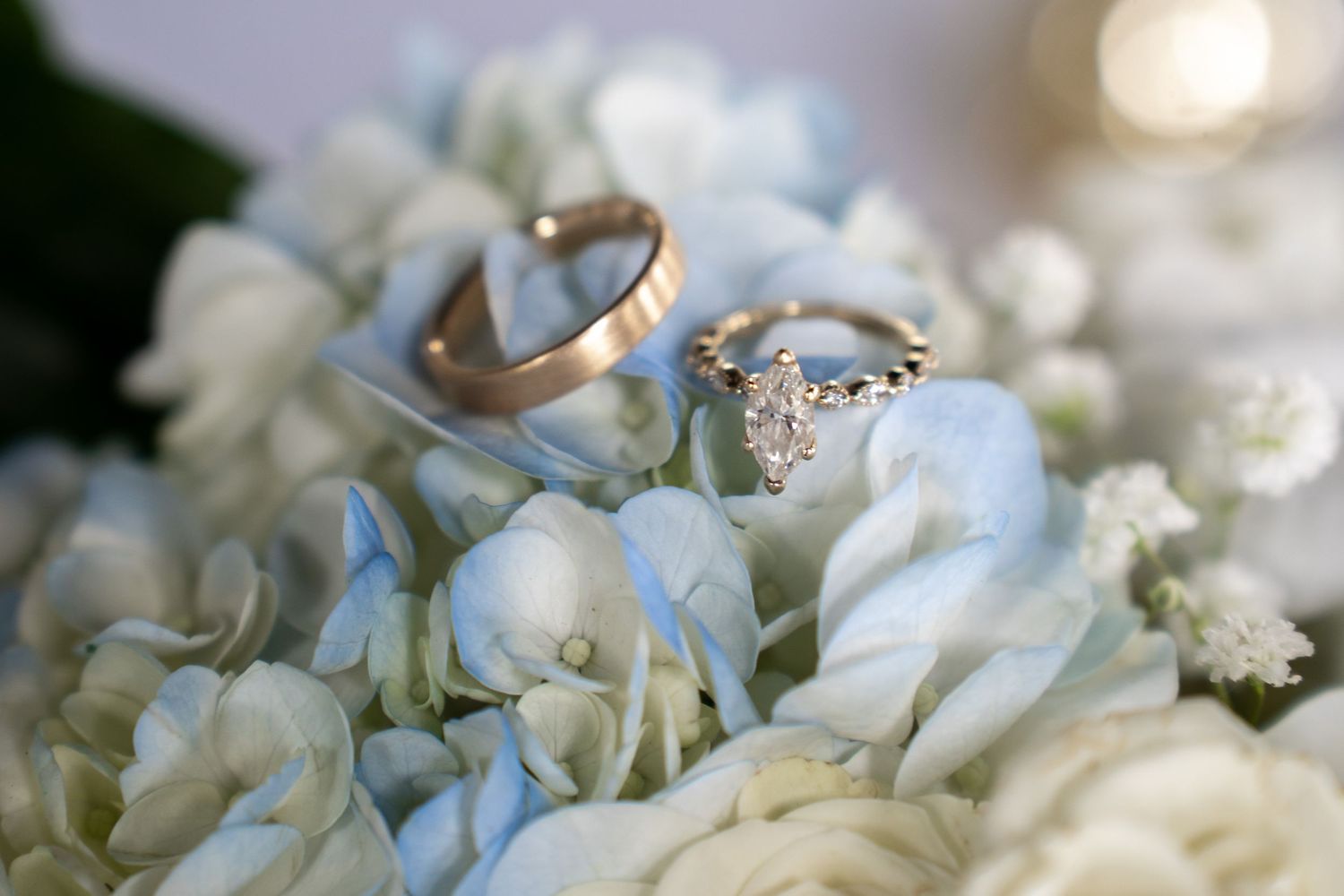 Two wedding rings, a gold band and diamond engagement ring, resting on soft blue hydrangea flowers.