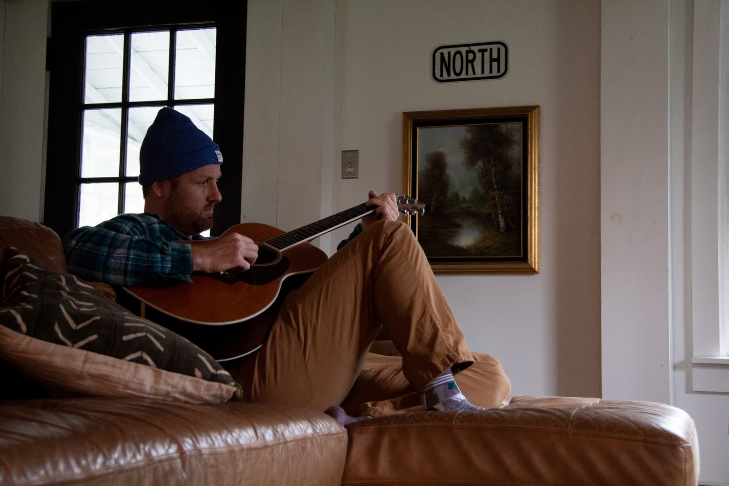 Man in blue beanie playing acoustic guitar while relaxing on a brown leather couch indoors.
