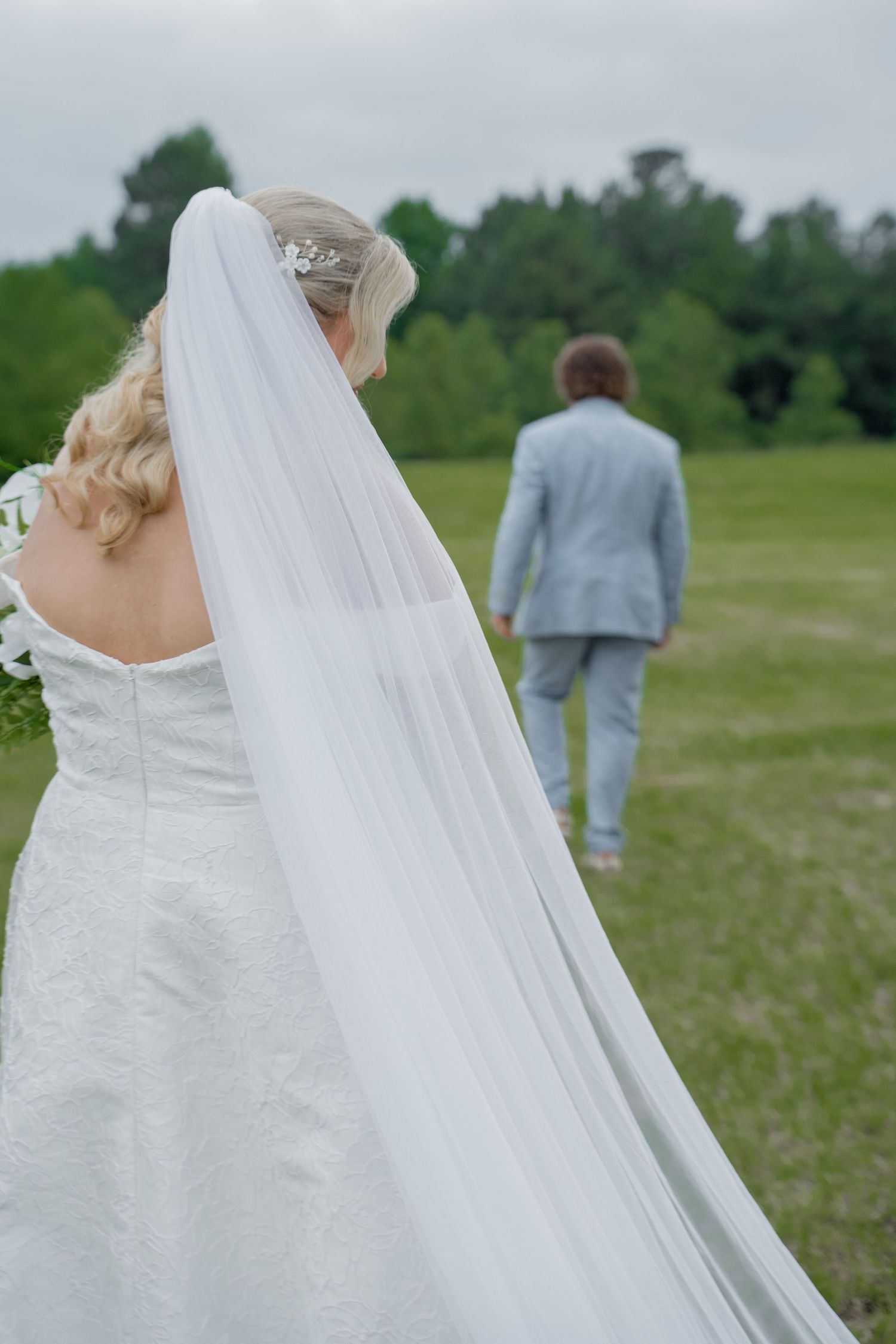 Bride in white gown and veil stands in green field as groom in light blue suit walks ahead.