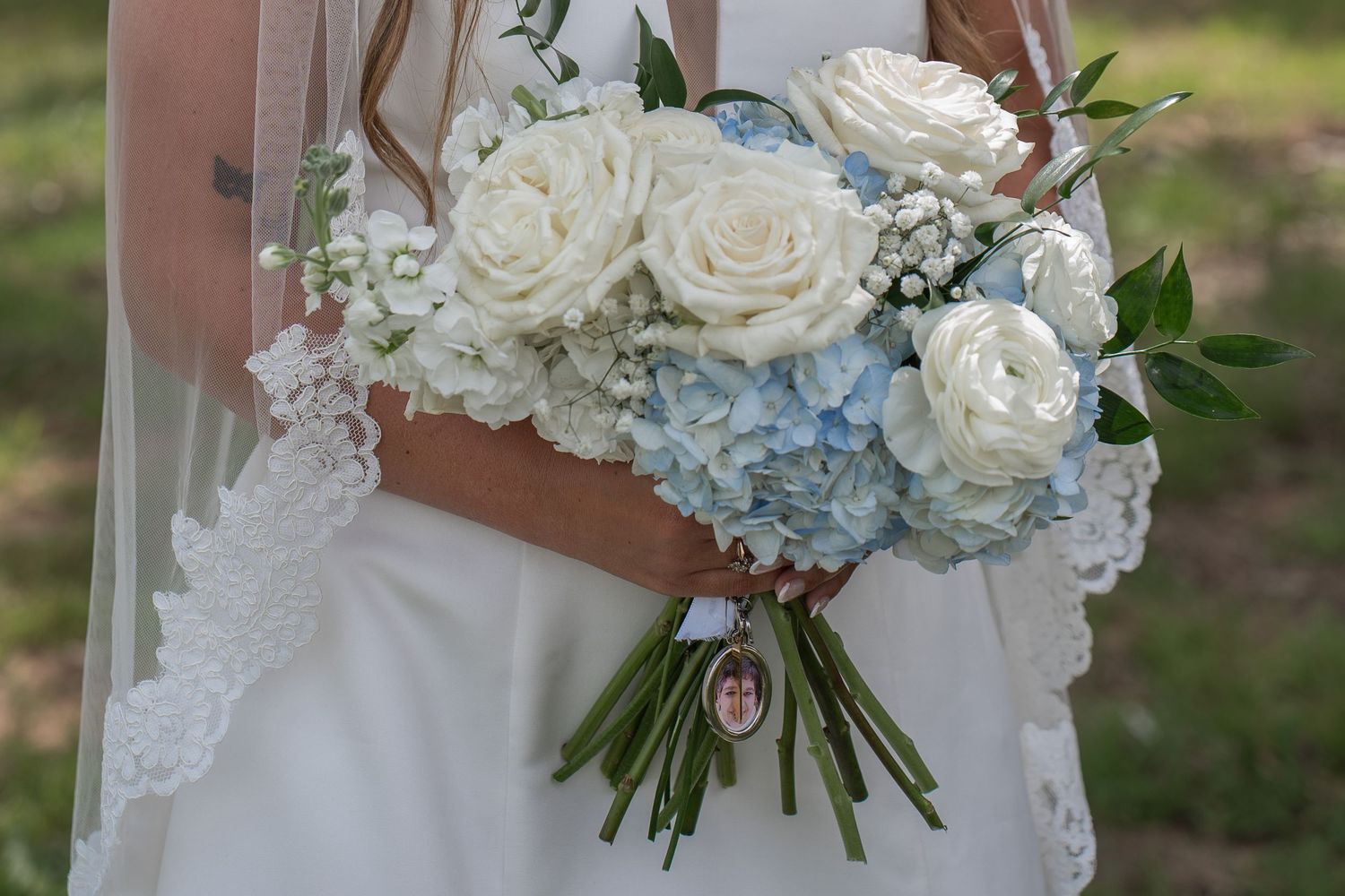 Bride holding a wedding bouquet of white roses and blue hydrangeas with a memorial charm on green stems.