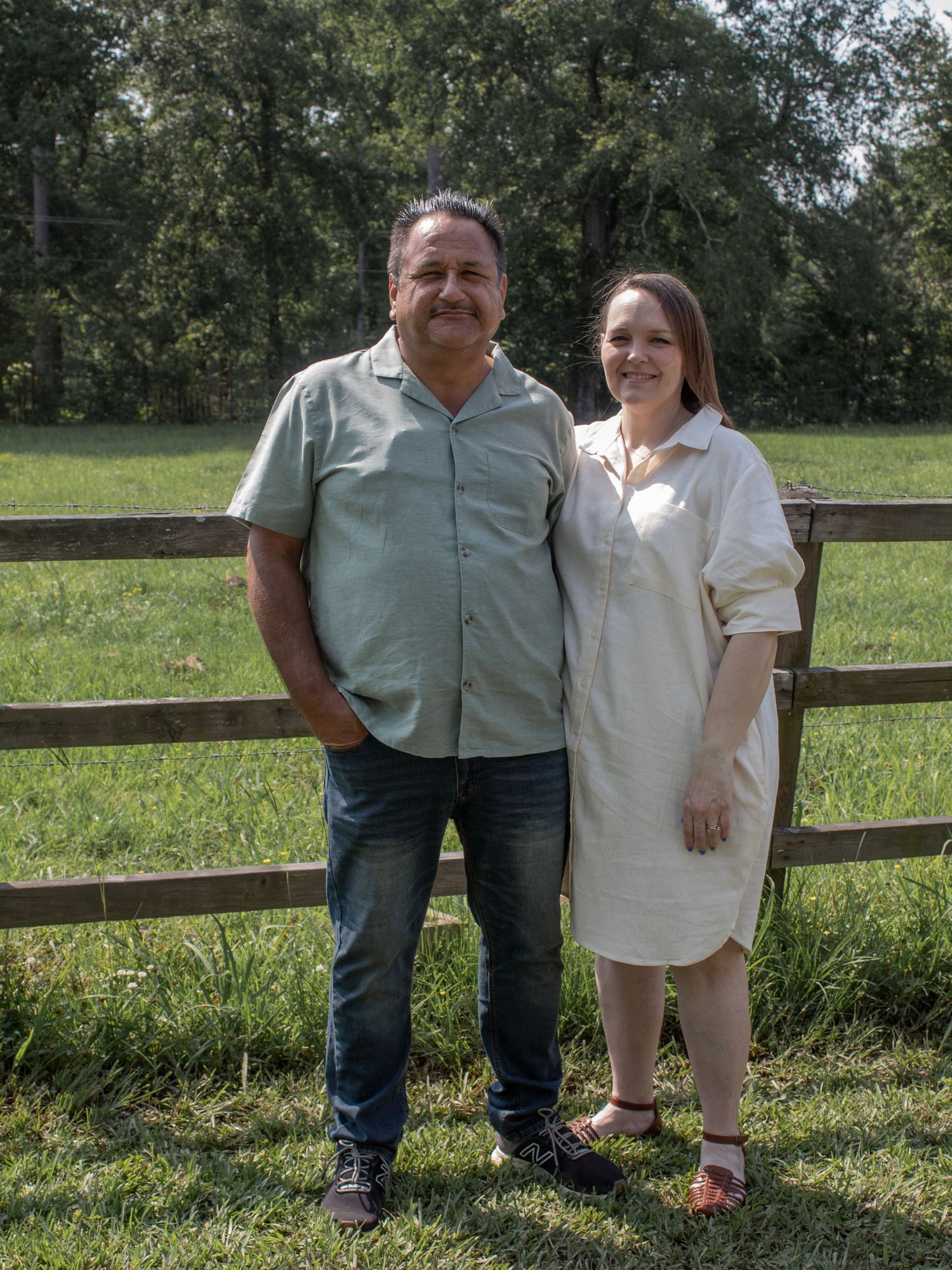 A man and woman smiling together outdoors by a wooden fence with green grass and trees in the background.