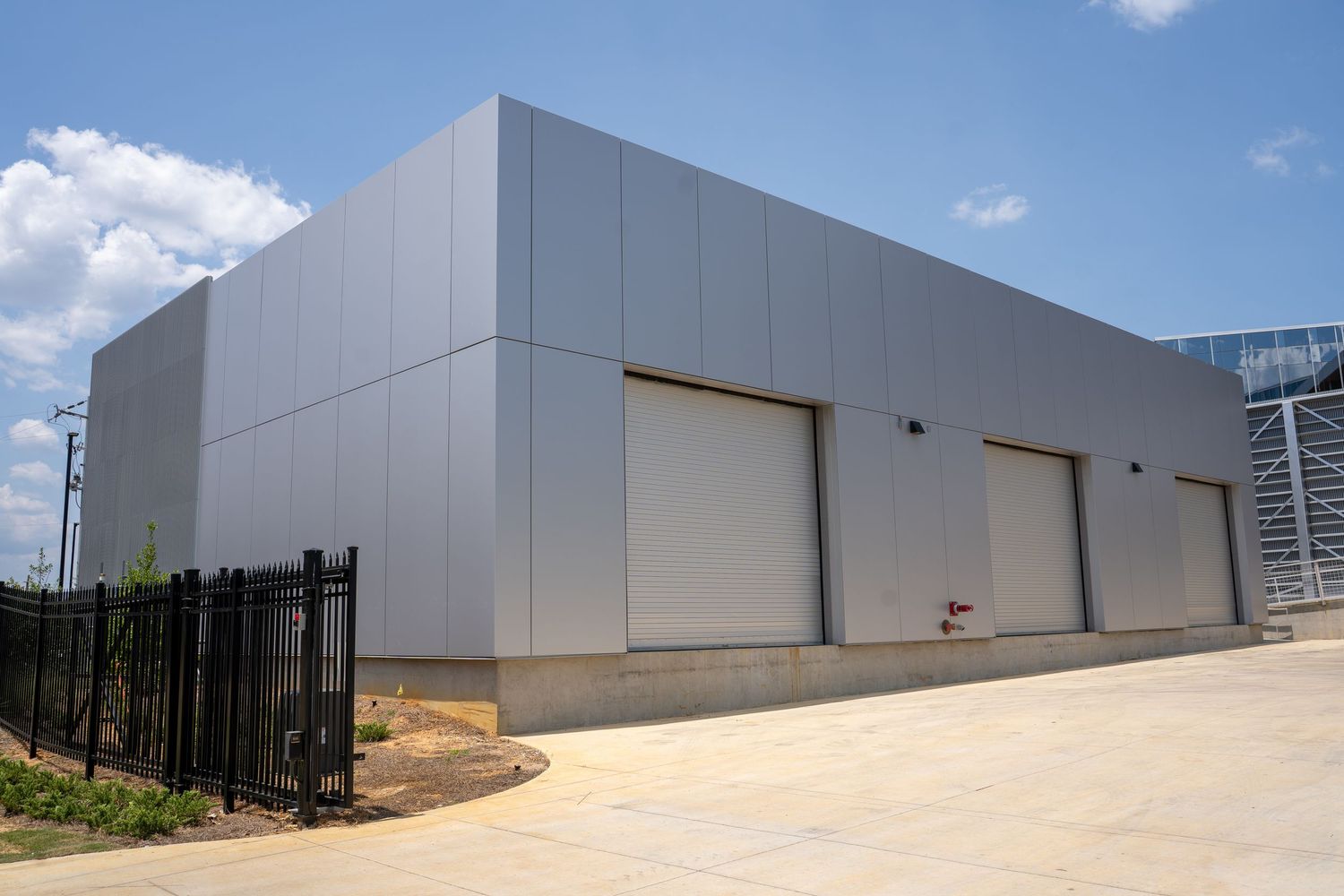 Modern gray metal industrial warehouse building with three roll-up garage doors and concrete driveway under blue sky.