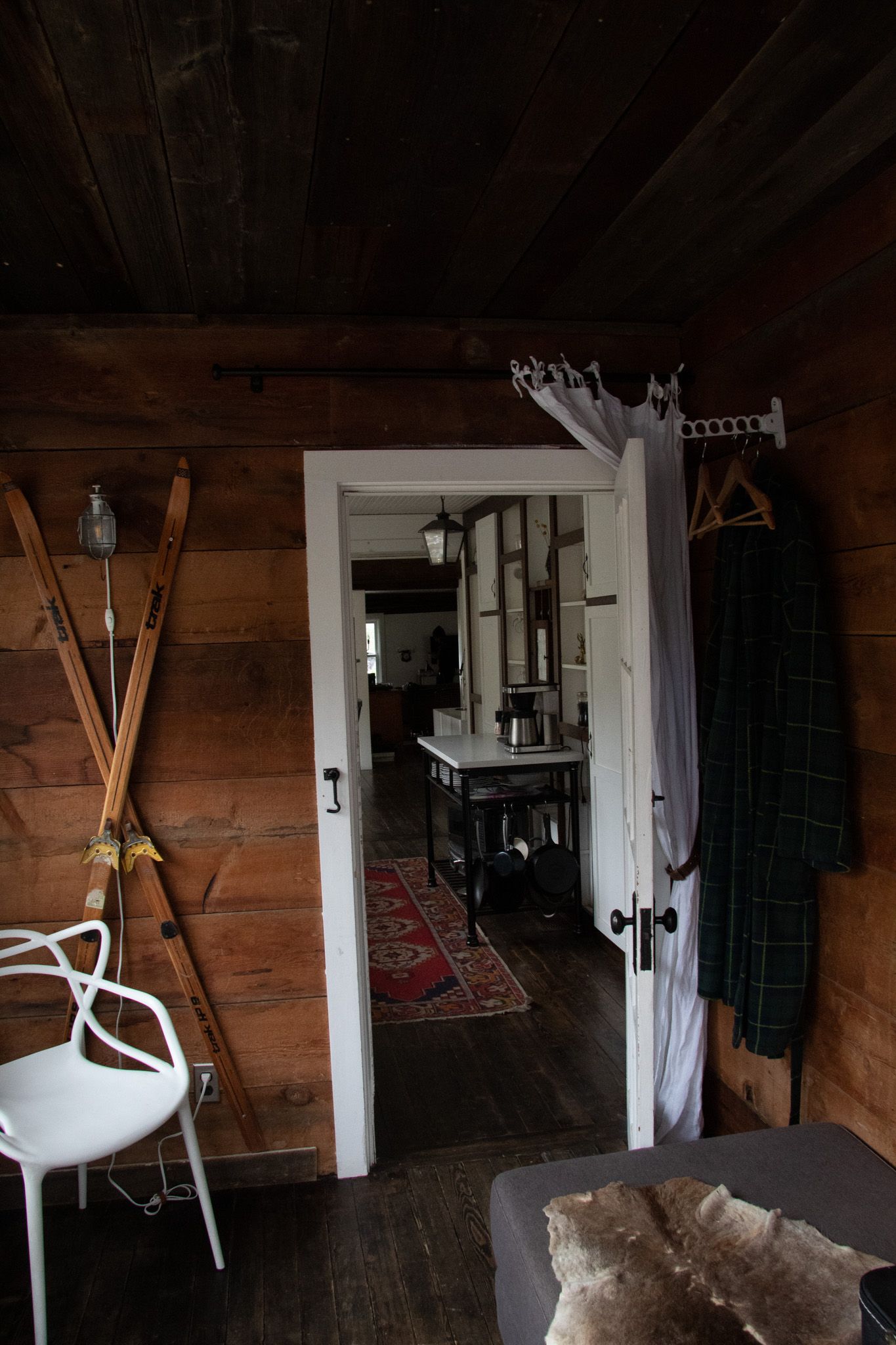 Rustic wooden cabin interior with vintage skis, white chair, open door revealing hallway with red rug, and hanging plaid jacket.
