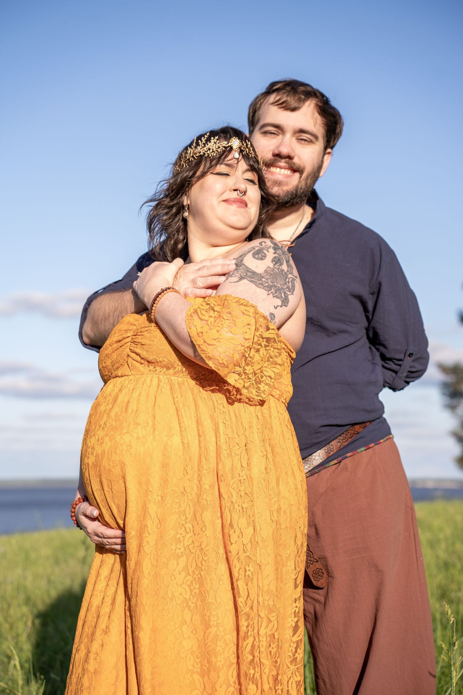 Pregnant woman in yellow lace dress with gold headpiece embraced by bearded man outdoors near water.