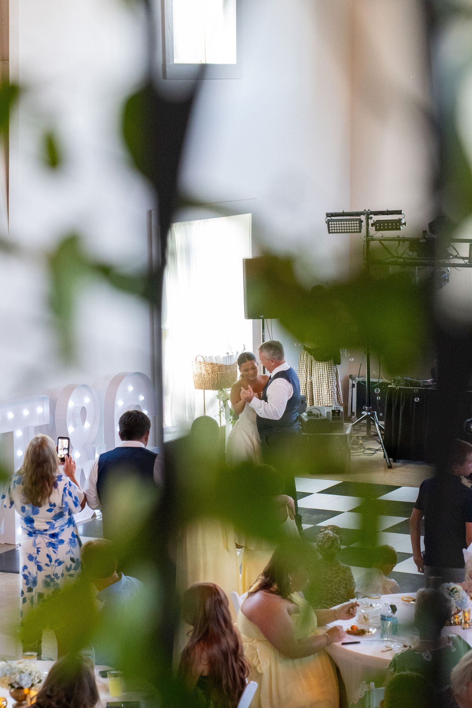 Bride and groom share their first dance at a wedding reception, viewed through green foliage with guests watching.