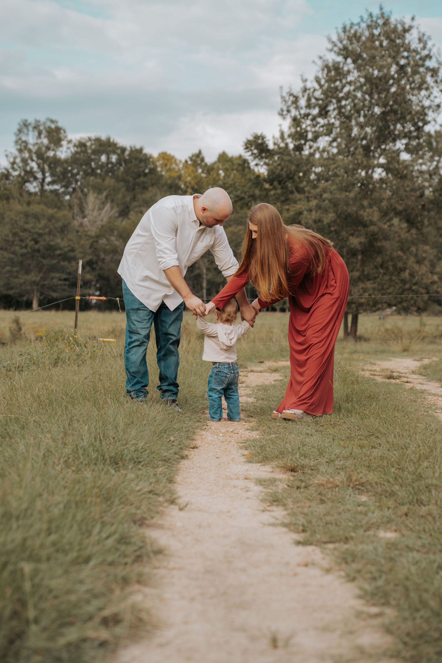 A family of three holds hands on a nature path, with parents in red and white leaning toward their toddler in jeans.