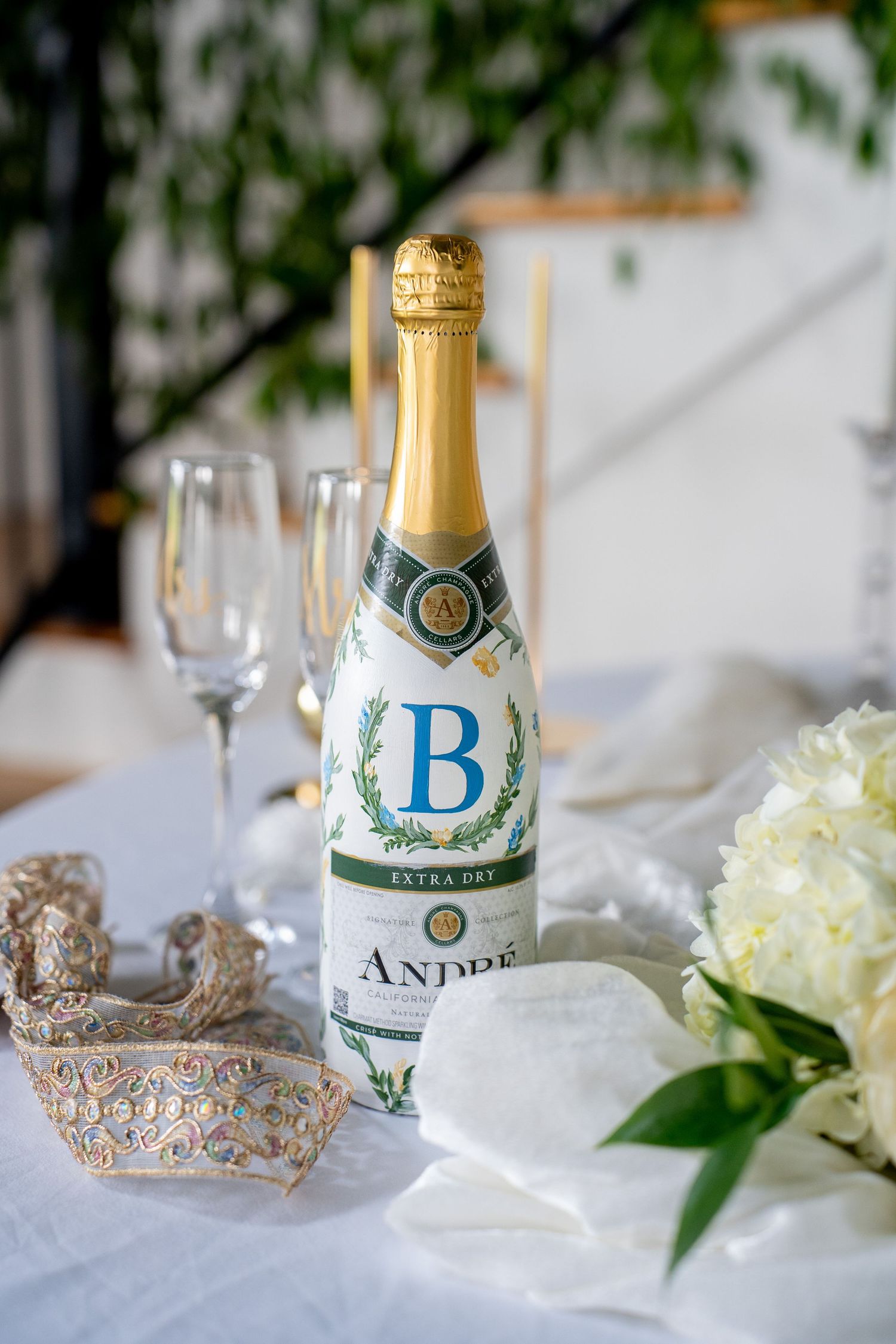 André Extra Dry champagne bottle with monogram B label beside crystal flutes and white flowers on elegant table.