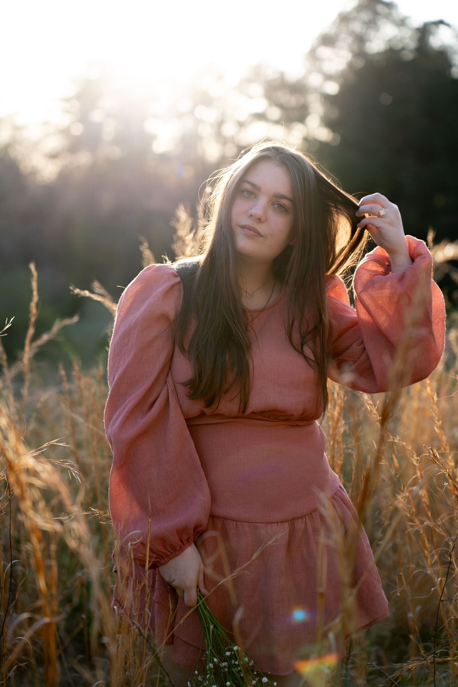 Young woman in pink dress standing in golden field at sunset, touching her long brown hair.