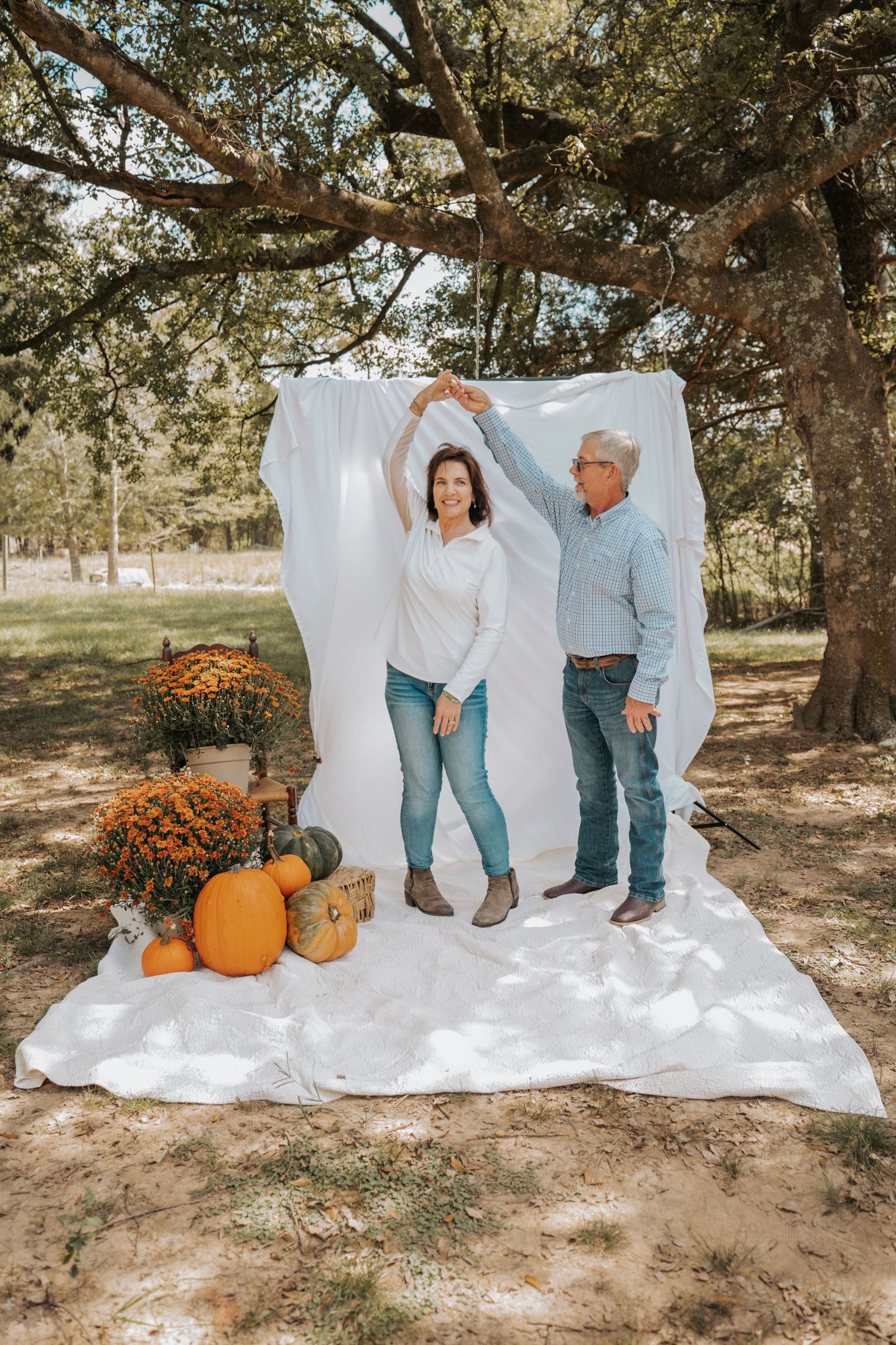 Couple dancing outdoors in fall setting with pumpkins and mums against white backdrop under large tree.