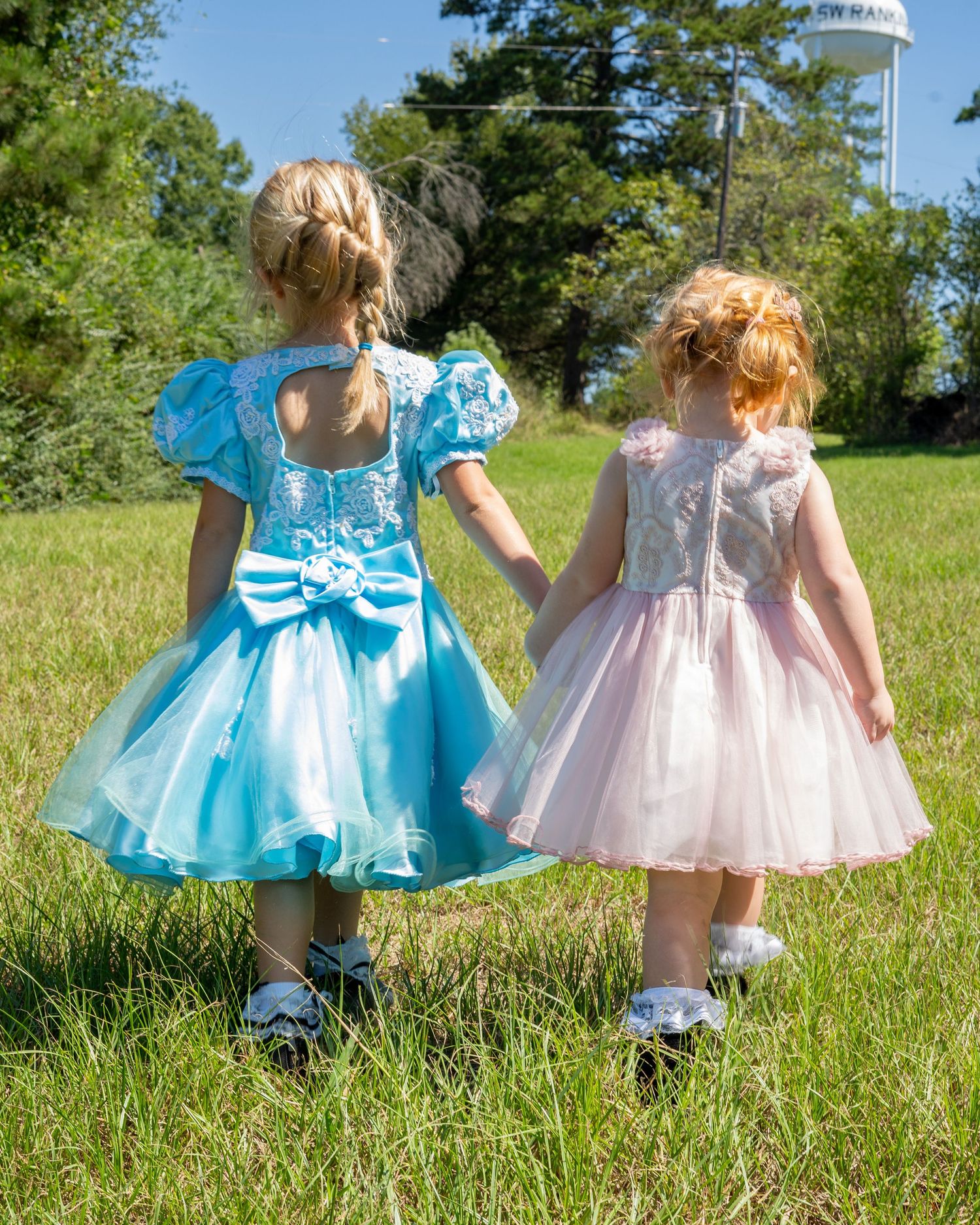 Two young girls in formal dresses holding hands walking through a grassy field on a sunny day.