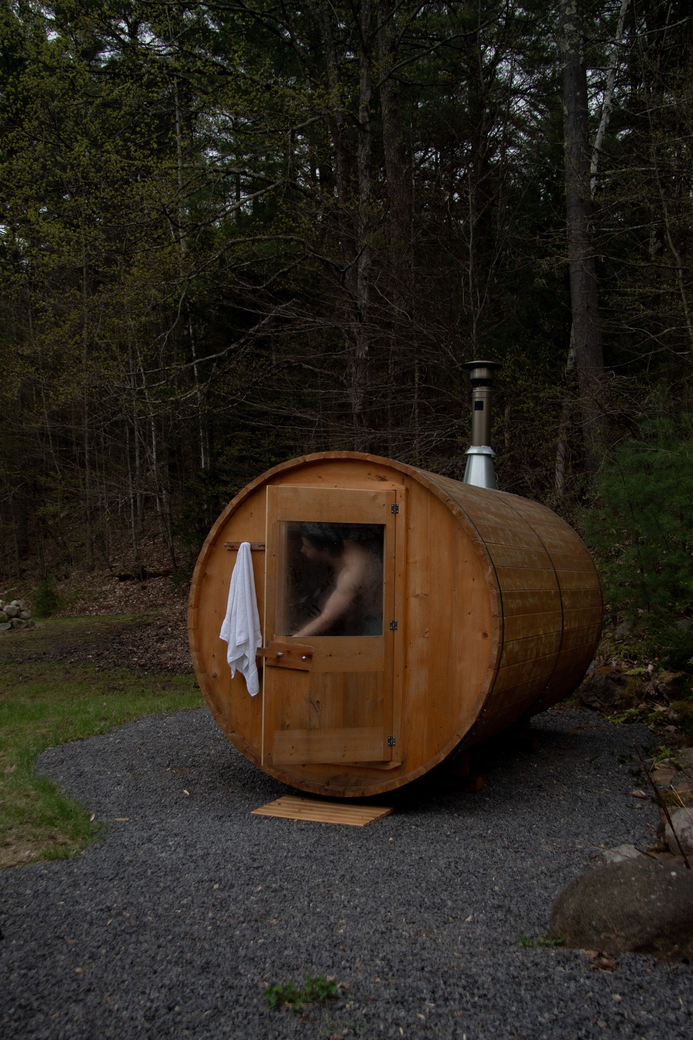 Barrel sauna with wooden door and white towel, set on gravel path surrounded by forest trees.