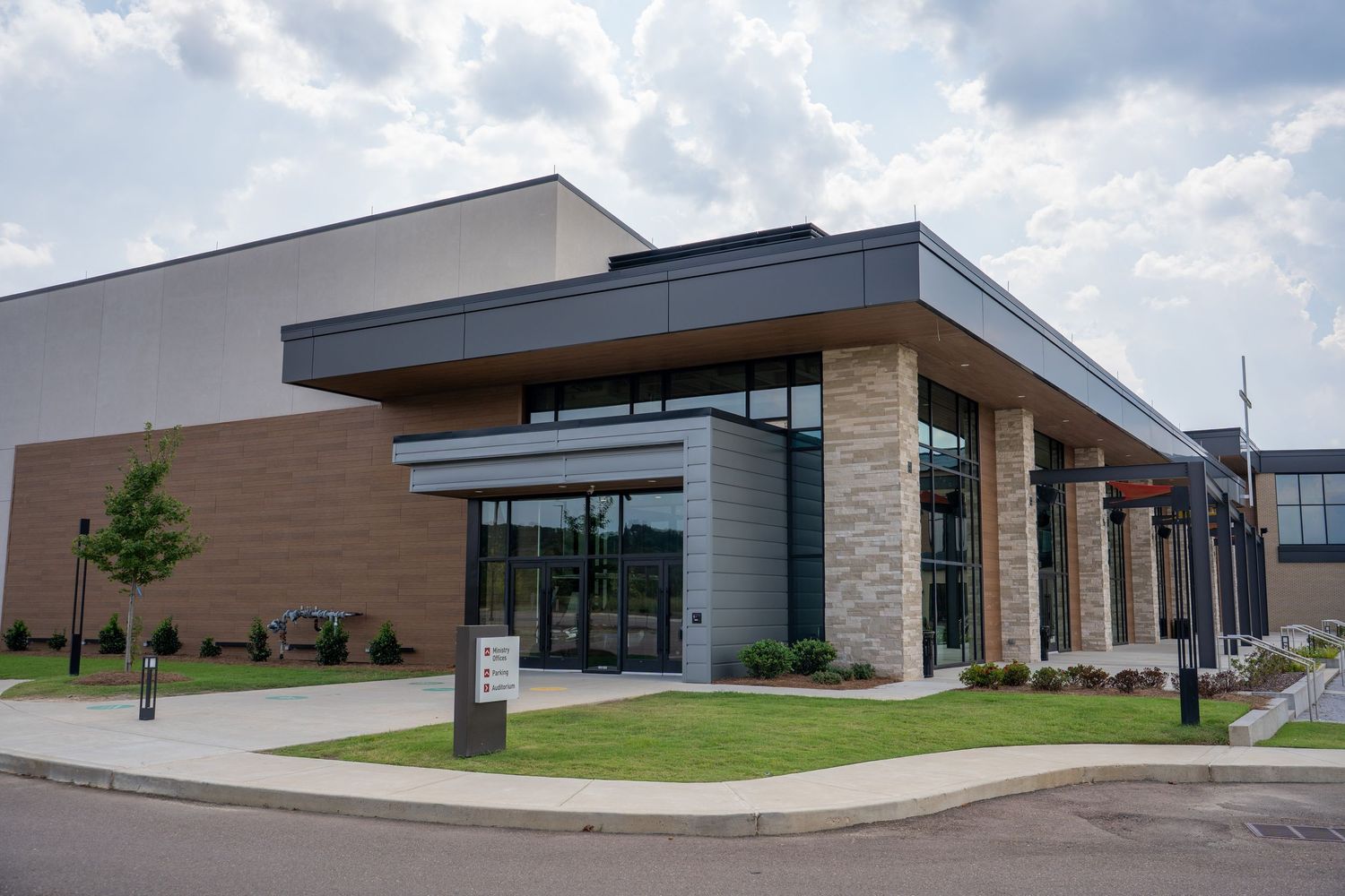Modern commercial building with brick and stone facade, large glass entrance, and manicured lawn under cloudy skies.