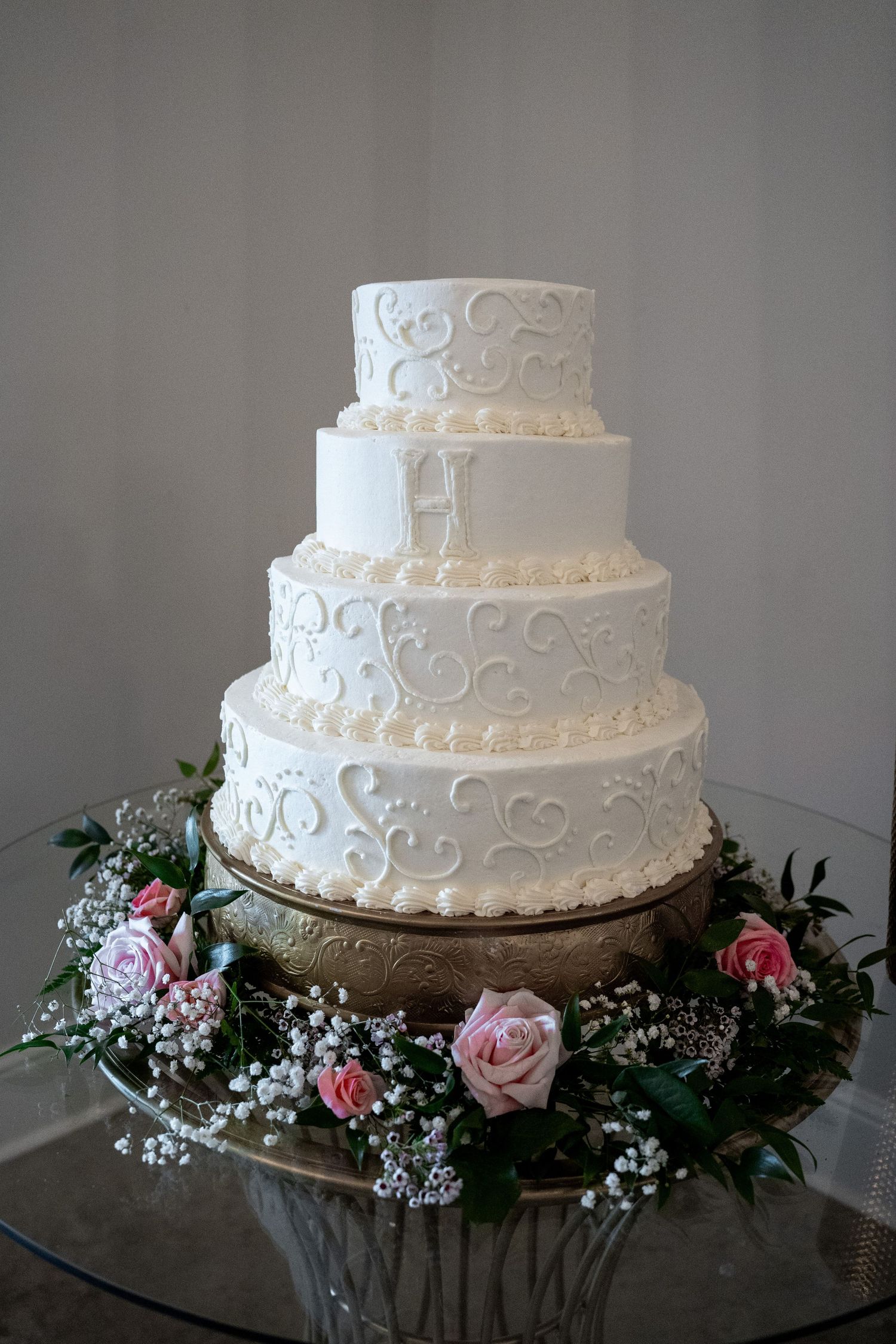 Elegant four-tier white wedding cake with scrollwork frosting, monogram, and pink roses on a gold stand.