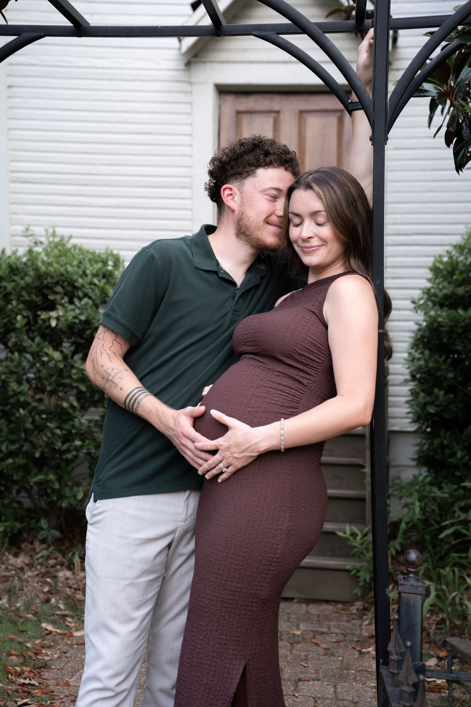 A couple tenderly embraces outdoors, the pregnant woman in a brown dress cradling her baby bump with her partner.