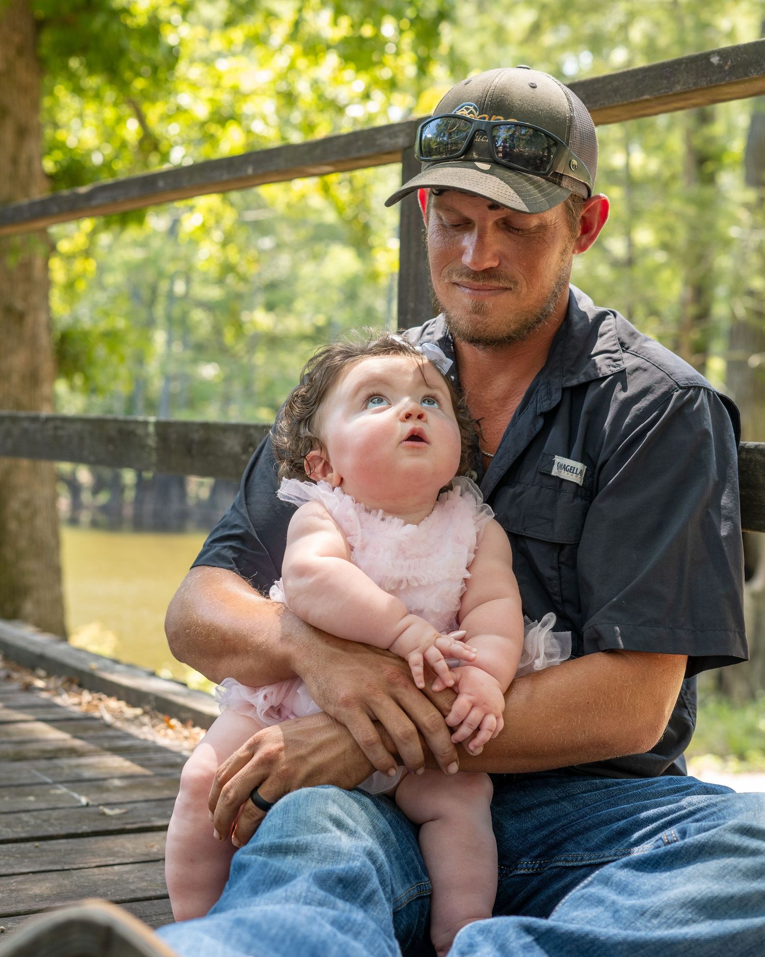 A man in a gray shirt and cap holds a baby girl in a pink dress on a wooden deck near a tree-lined pond.