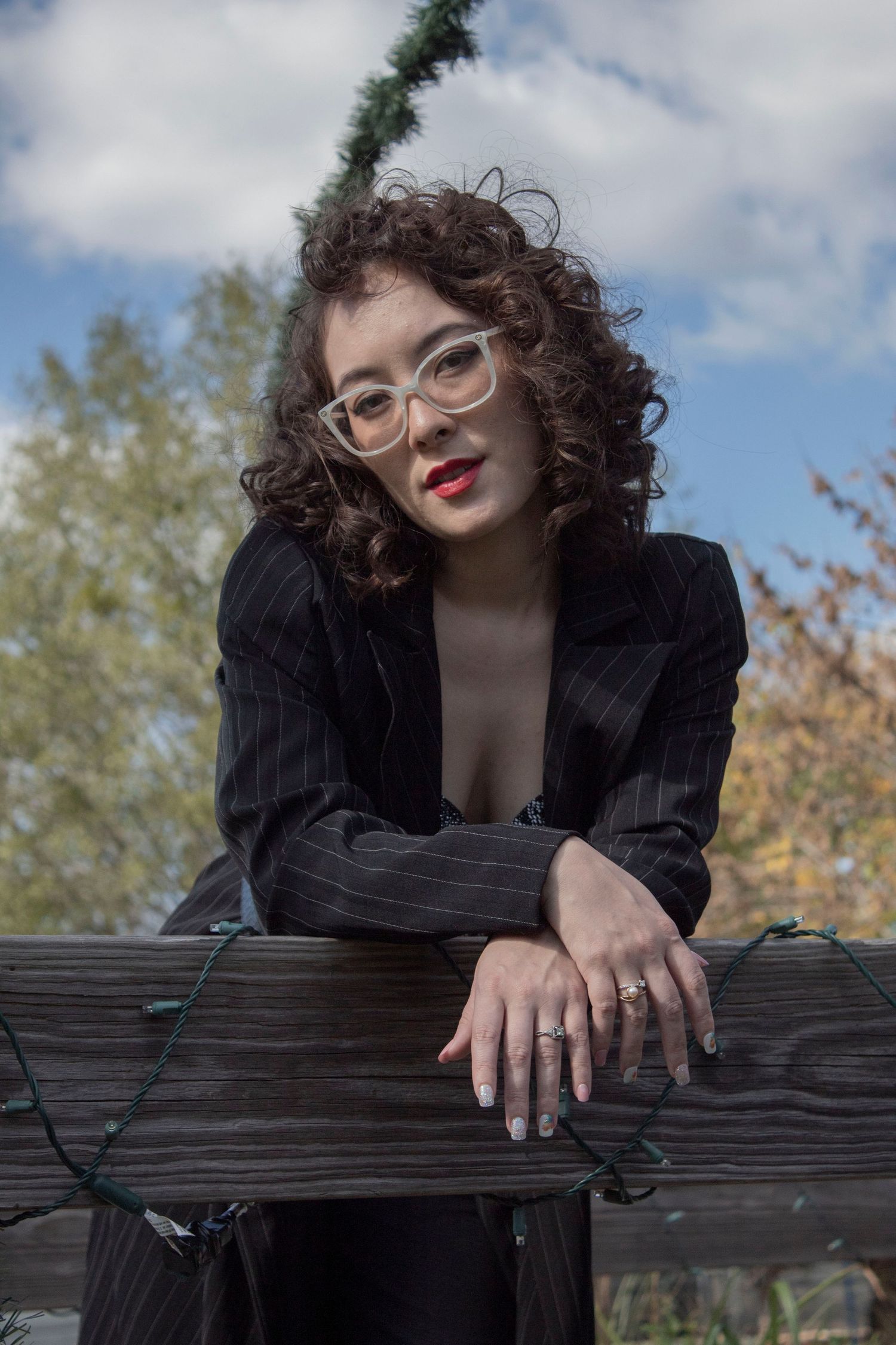Young woman with curly hair and glasses in black blazer leans on wooden fence outdoors on sunny day.