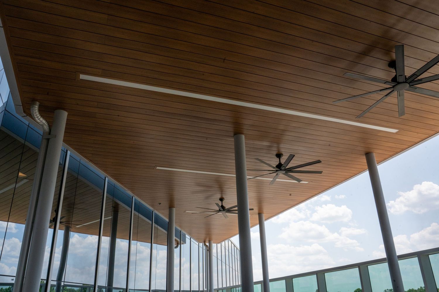 Modern covered patio with wood ceiling, ceiling fans, and floor-to-ceiling windows overlooking a scenic outdoor view.