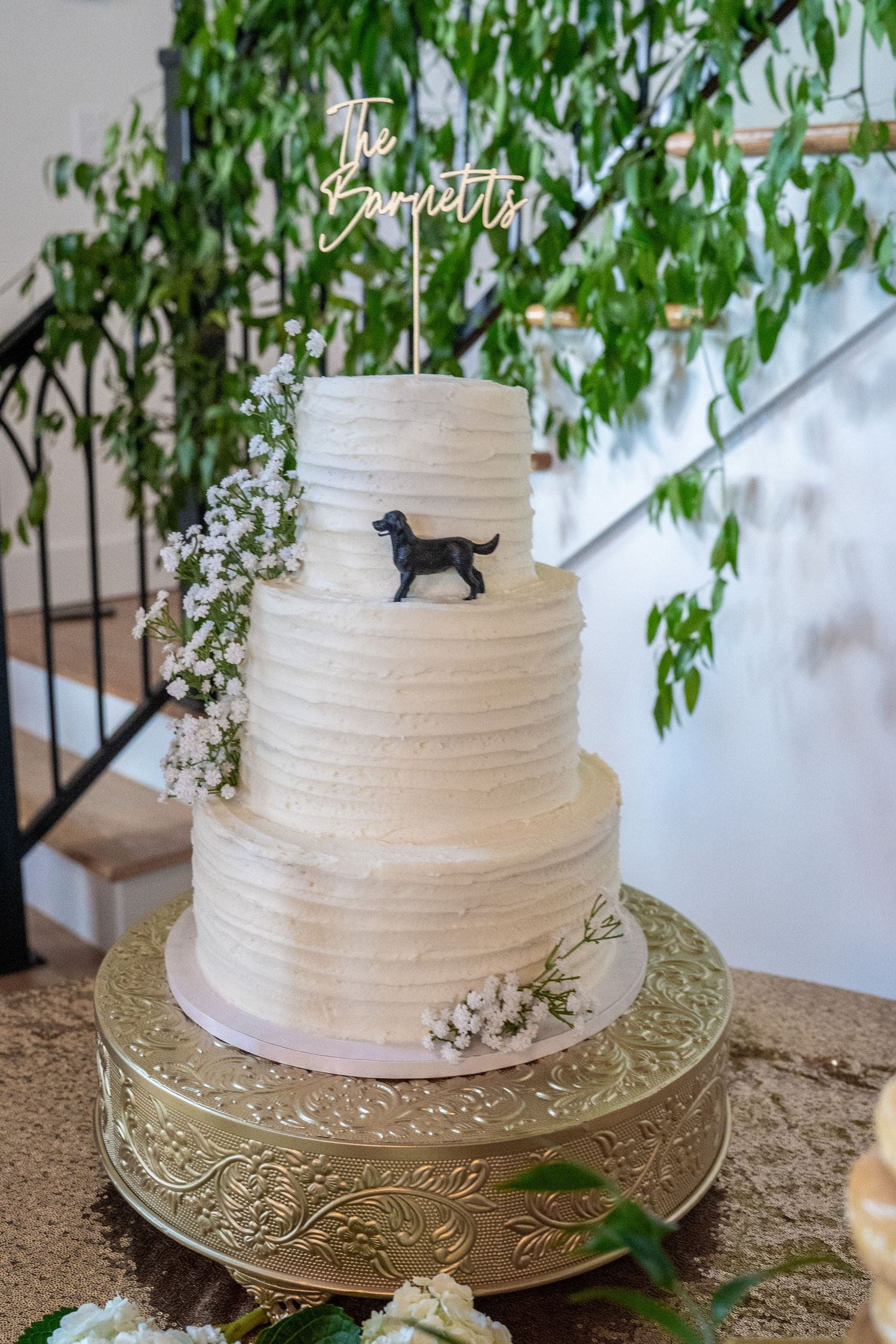 Three-tier white textured wedding cake with dog silhouette topper, baby's breath flowers, on gold stand.