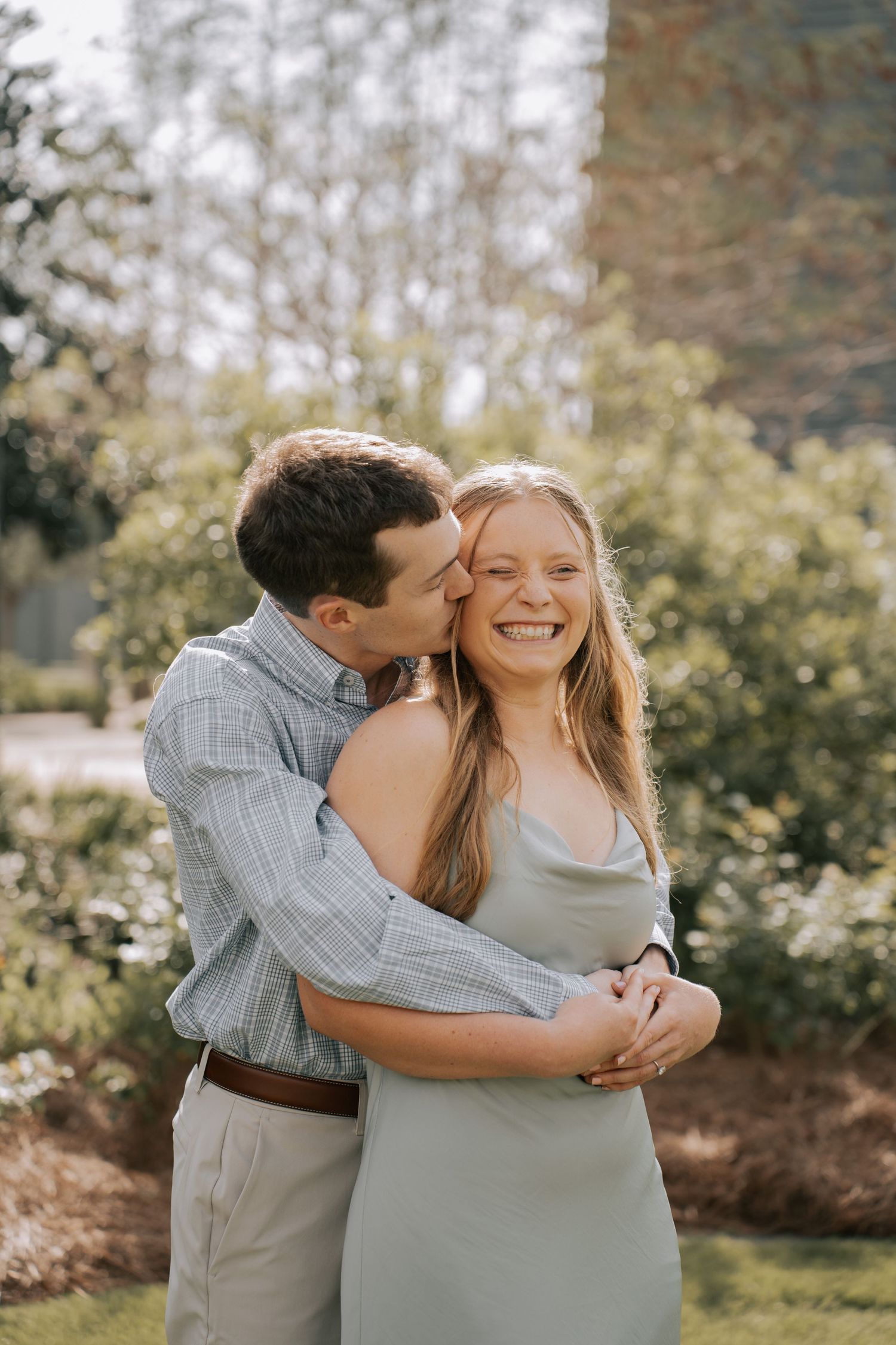 A happy couple embraces outdoors, man kissing woman on cheek as she smiles, surrounded by lush green bushes.