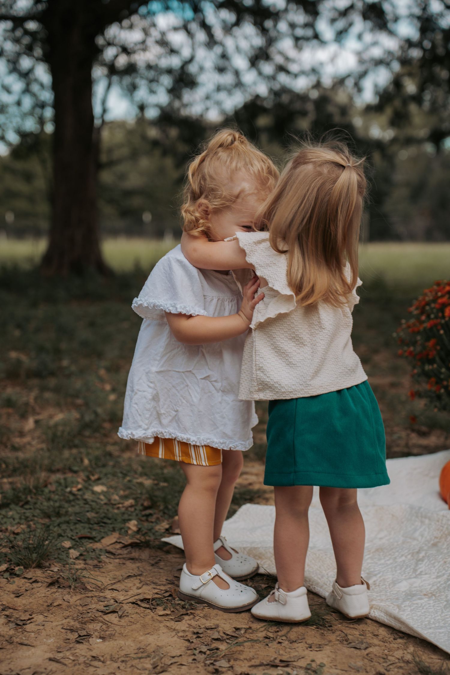 Two young toddler girls in white tops hugging outdoors in a park setting with green trees in the background.
