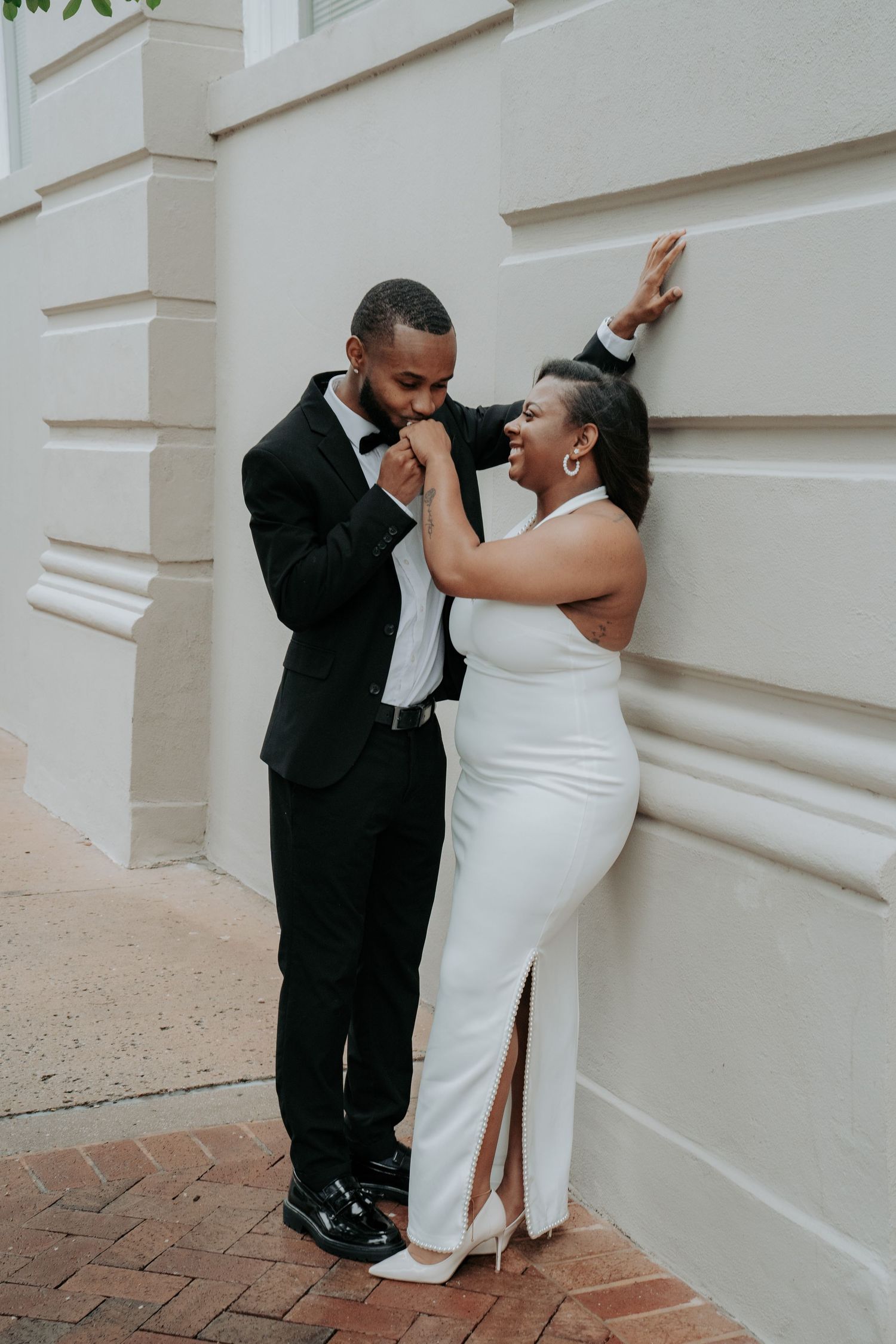 A couple poses romantically against a wall, man in black suit kissing womans hand, woman in white halter gown smiling.