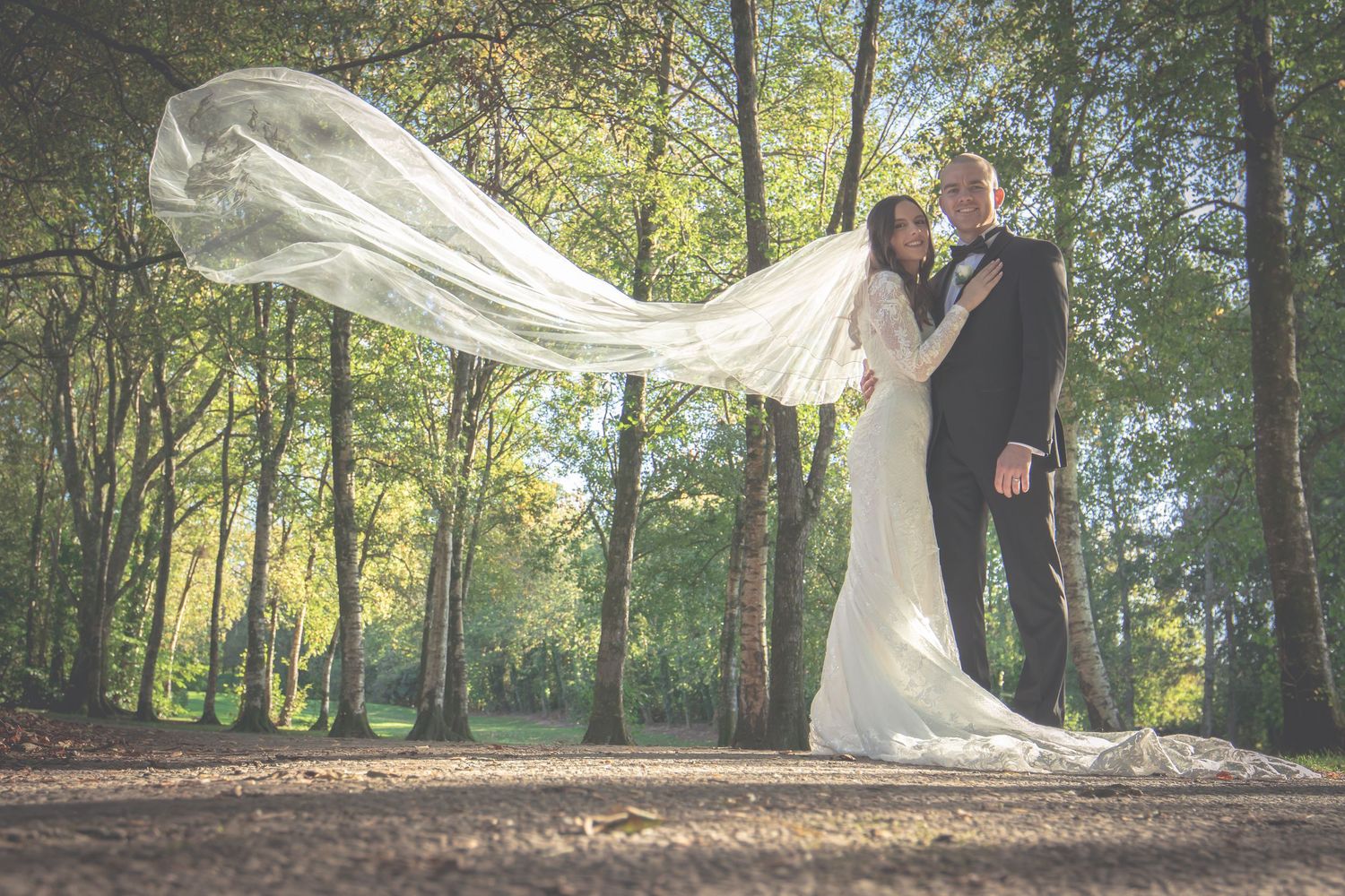Bride and groom embracing in woodland with dramatic veil flowing in the wind.
