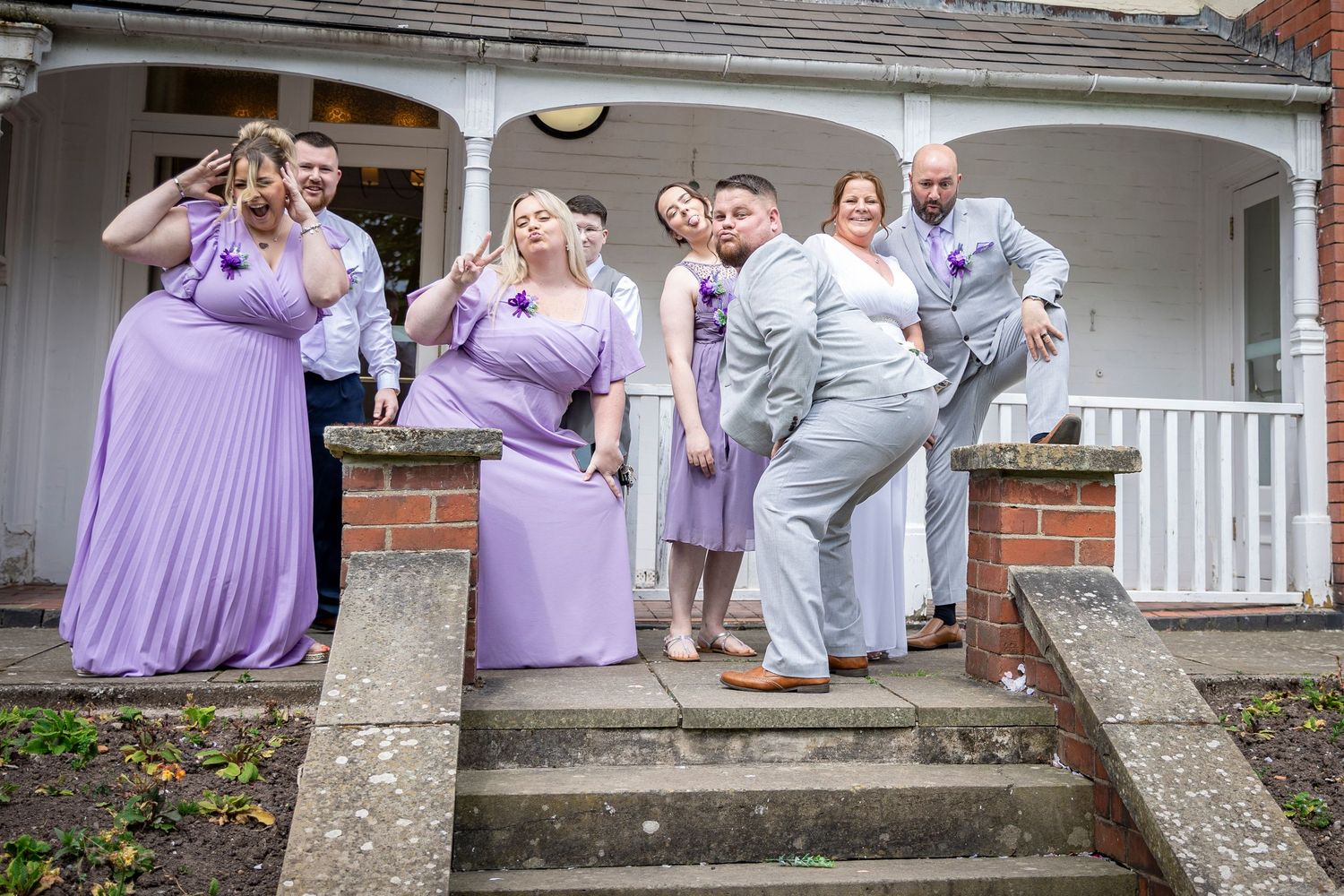 Bridal party in purple dresses and grey suits striking fun poses on steps.