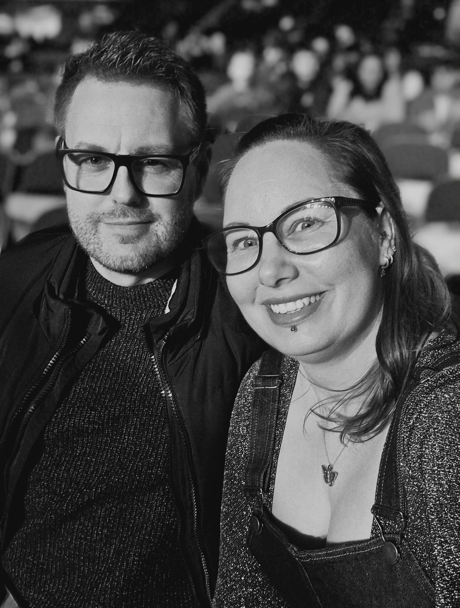 Black and white photo of a smiling couple wearing glasses posing together at an indoor event.