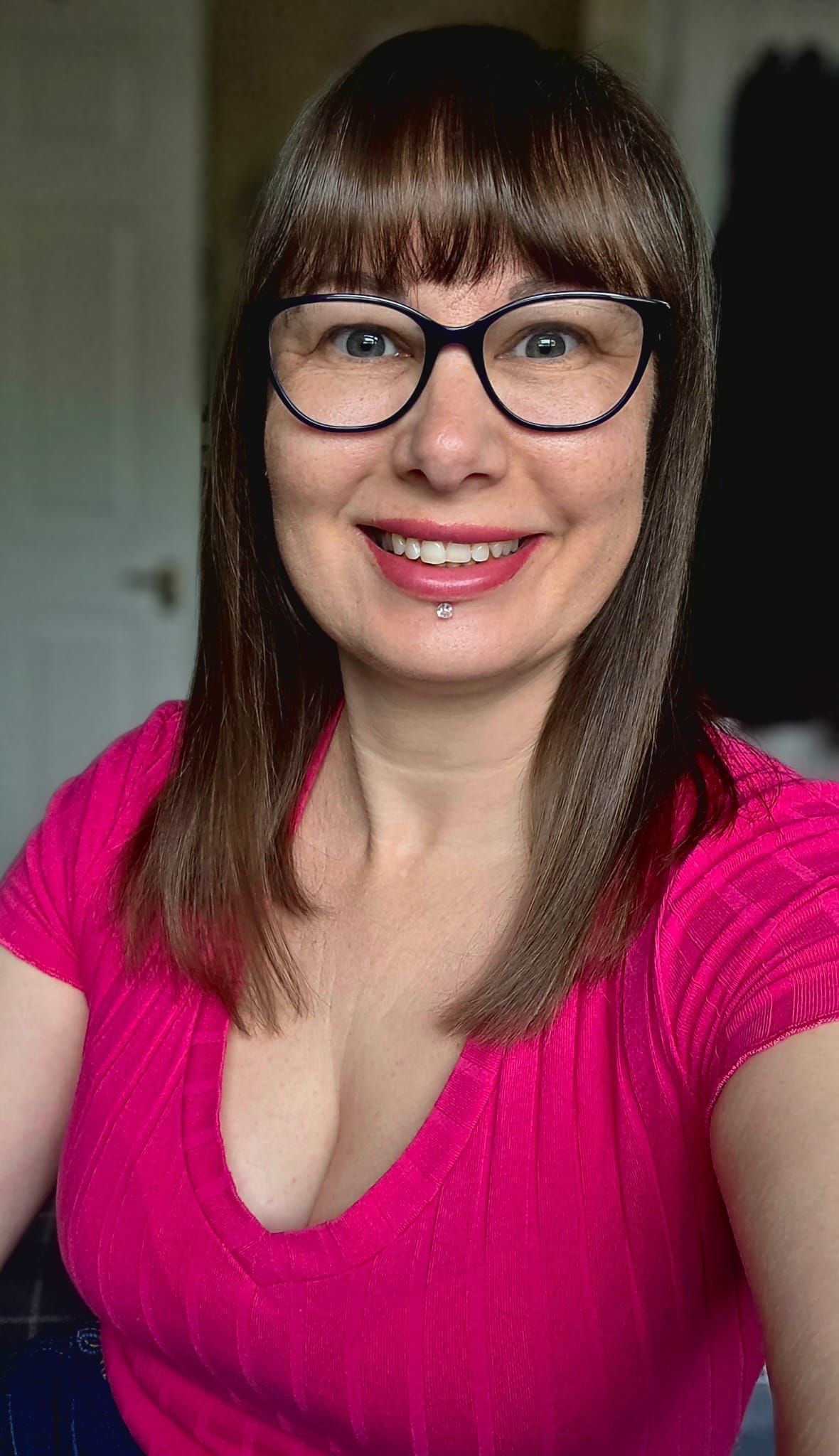Smiling woman with brown hair, bangs, and glasses wearing a pink top taking a selfie.