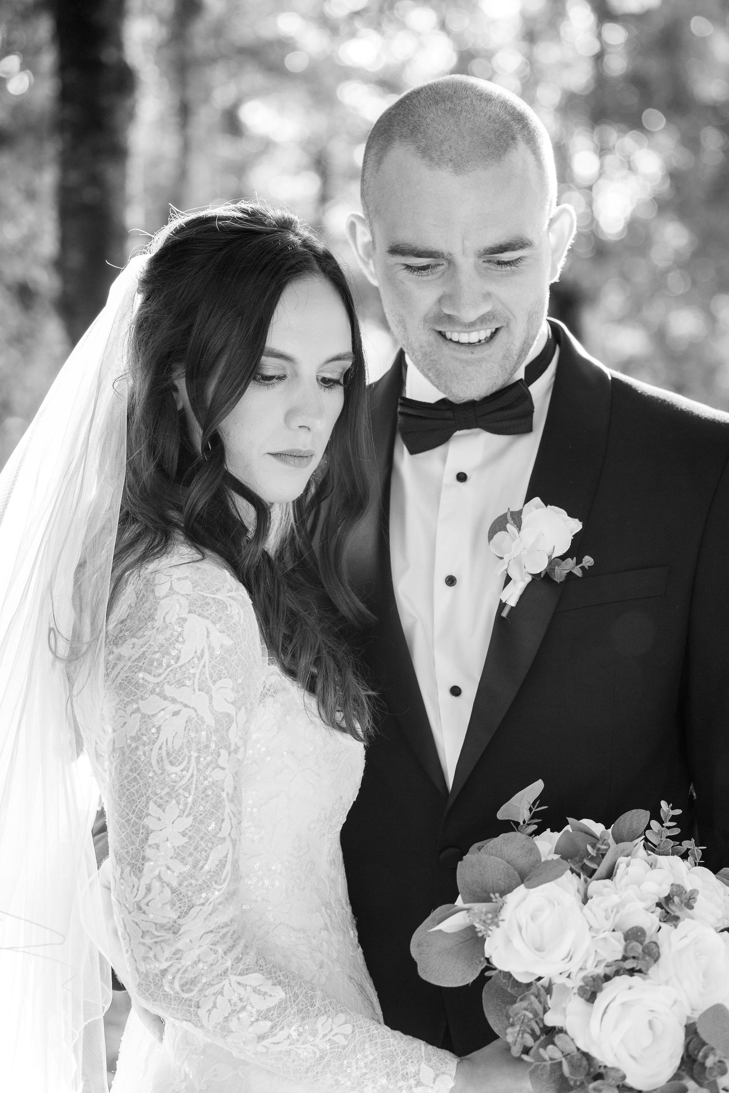 Black and white wedding portrait of bride in lace gown and groom in tuxedo holding bouquet of white roses outdoors.