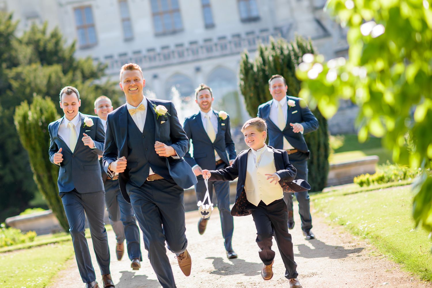 Groomsmen in navy suits running playfully across mansion lawn.
