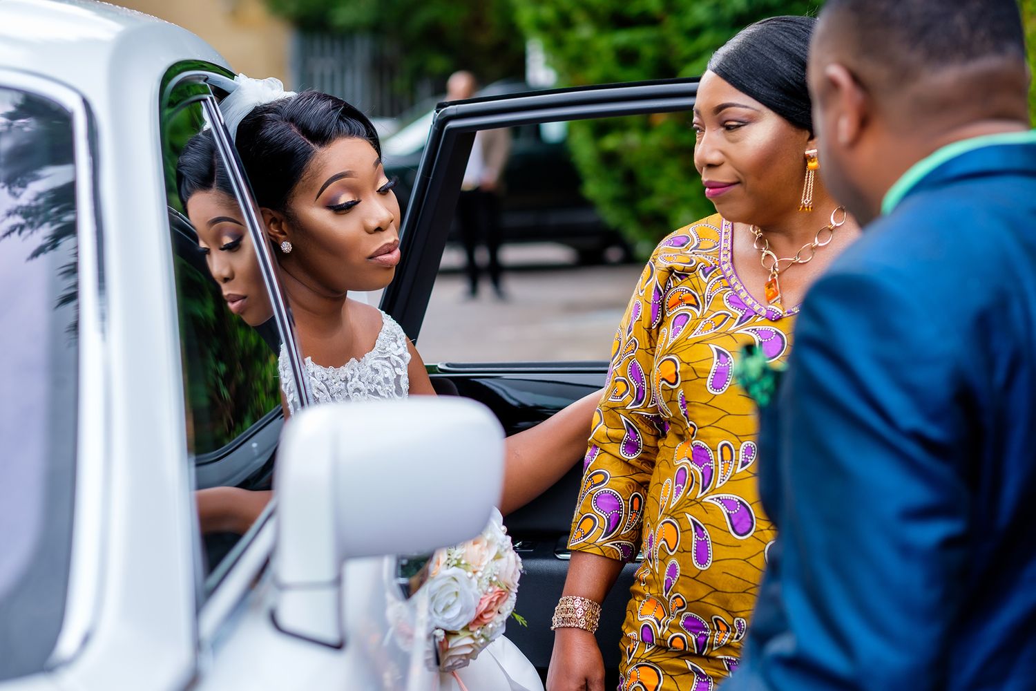 Wedding guests in traditional African prints and formal wear gather near cars.
