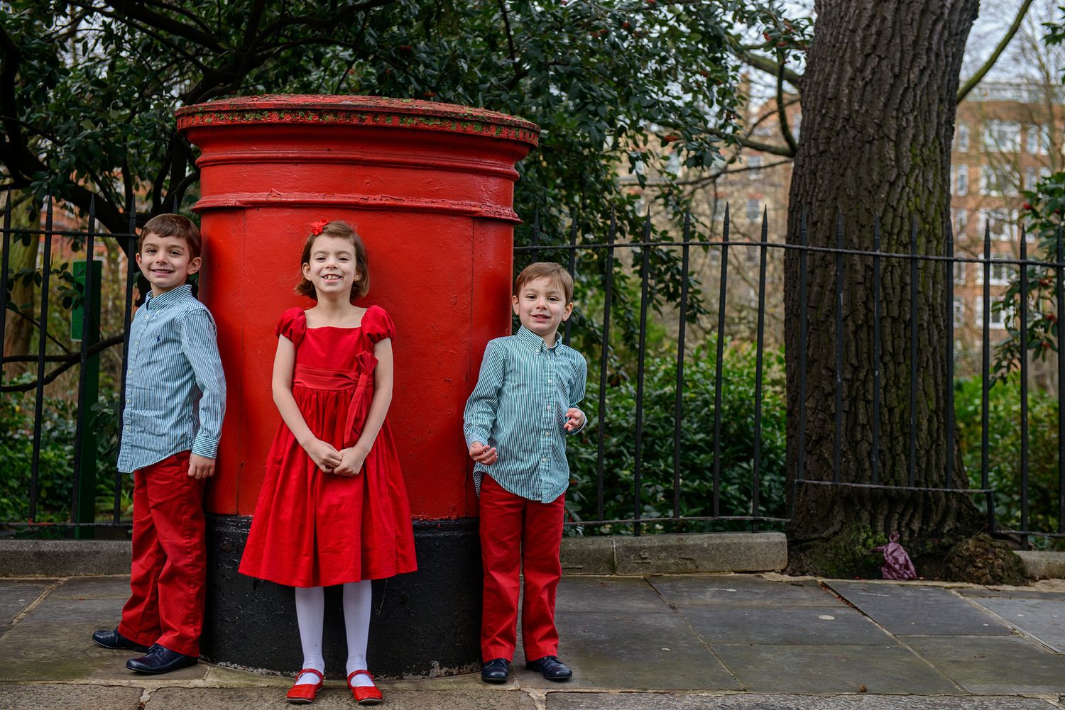 Children in red and blue outfits pose near a bright red British post box on a sunny day.