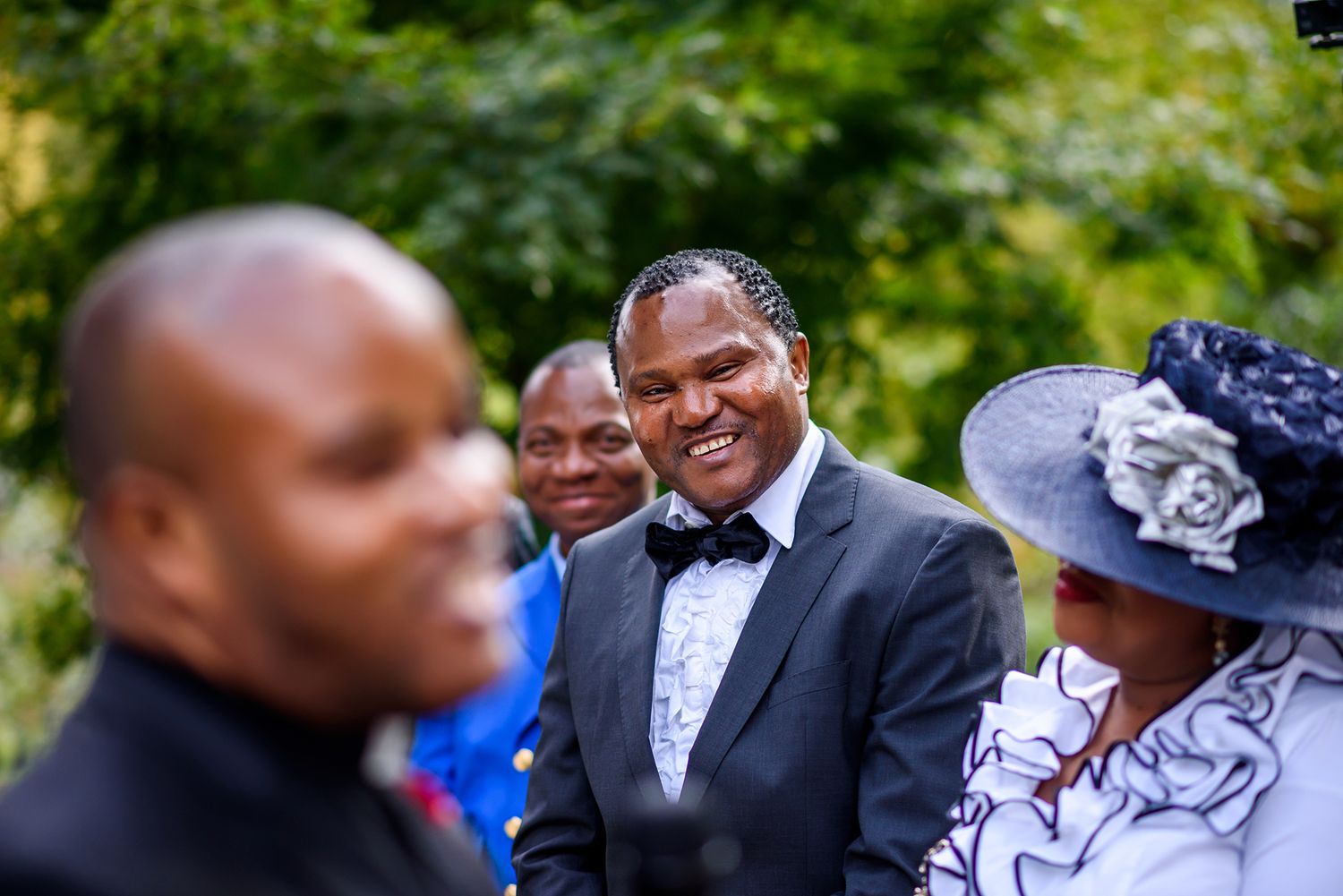 Wedding guests and celebrants gathered outdoors on a sunny day with lush green foliage.