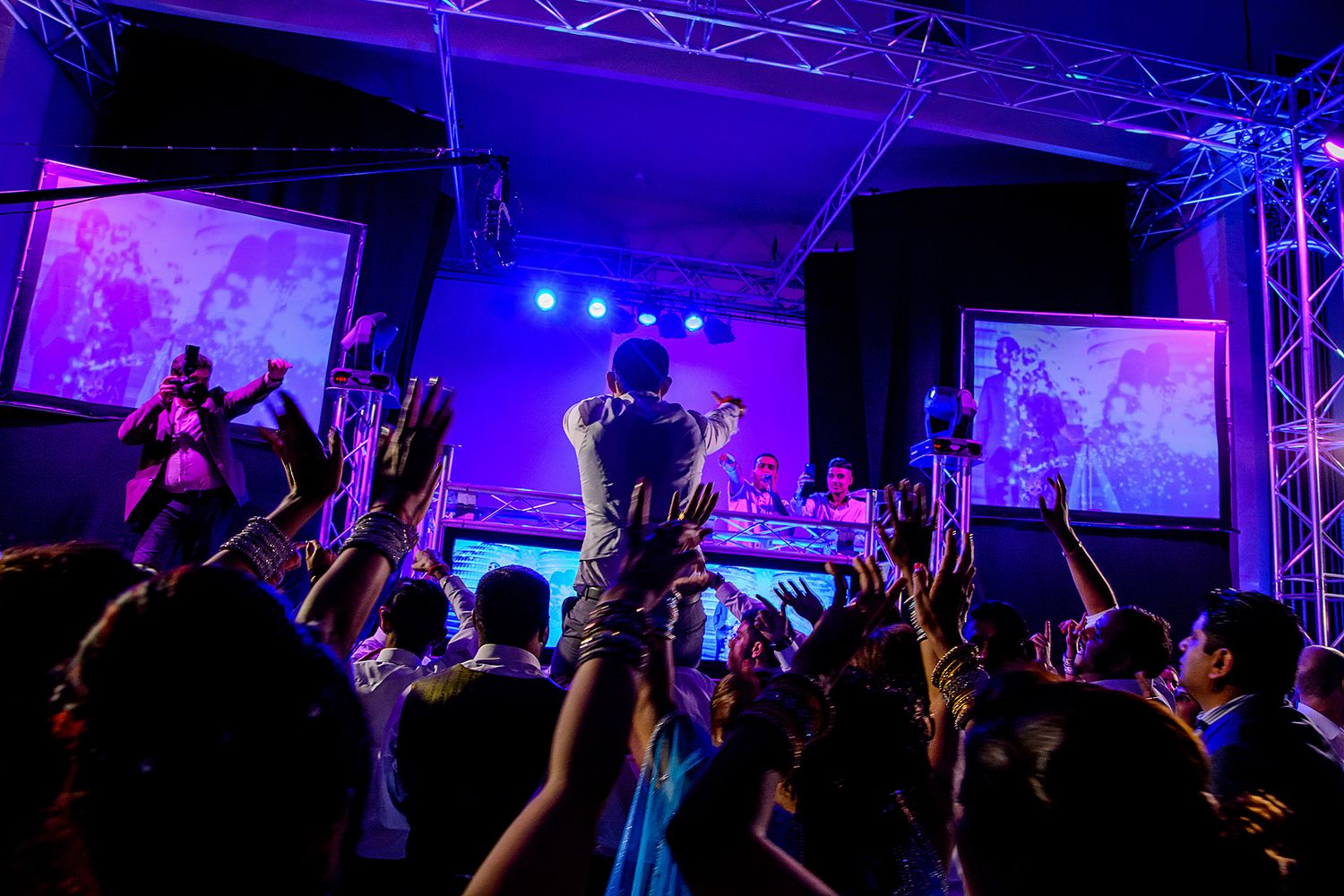 Concert crowd silhouetted against purple and blue stage lights with raised hands at a music event.