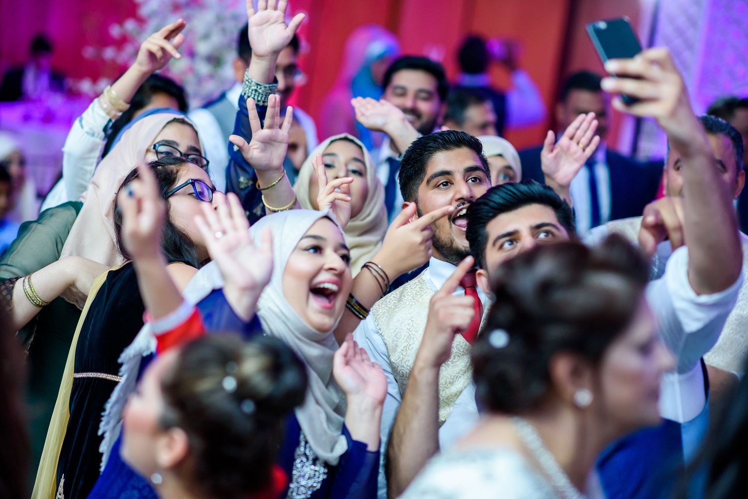 Enthusiastic wedding guests dancing and celebrating with raised hands under purple lighting.