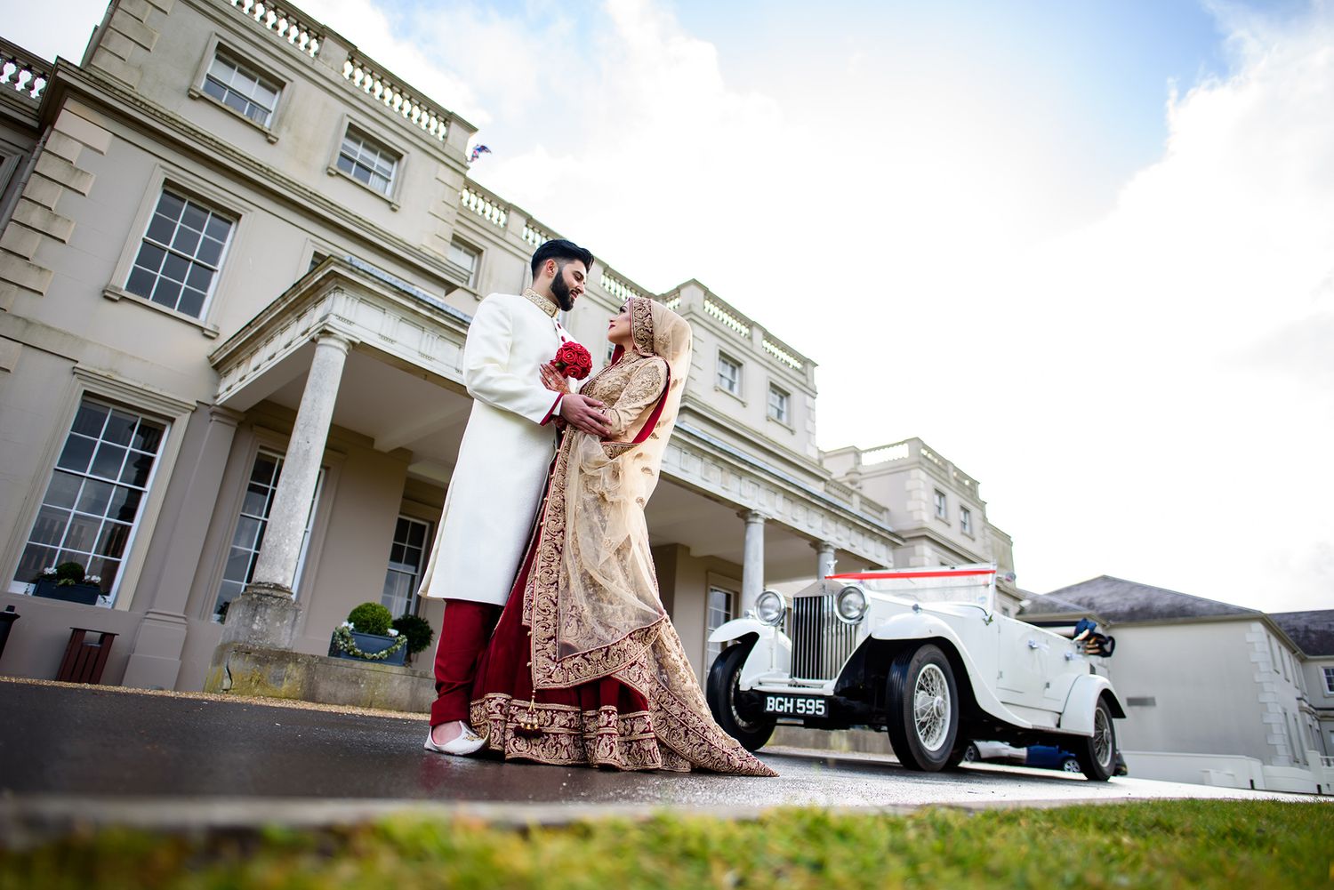 A couple in traditional attire pose in front of a grand mansion with a vintage car.