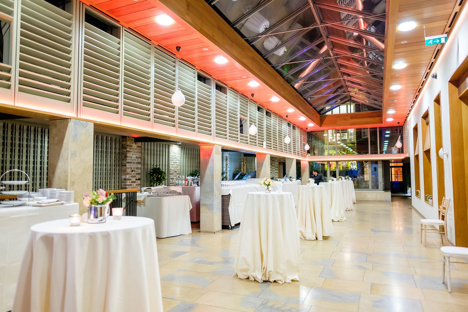 Large empty event venue hall with white round tables setup under orange lighting and high ceilings.