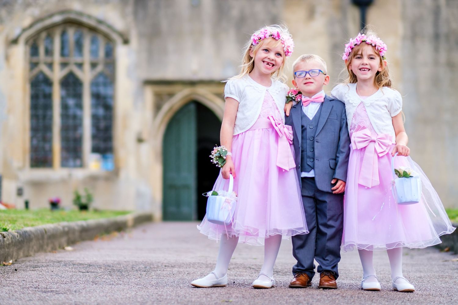 Three children in formal attire with pink dresses and flower crowns stand together outside a historic church entrance.