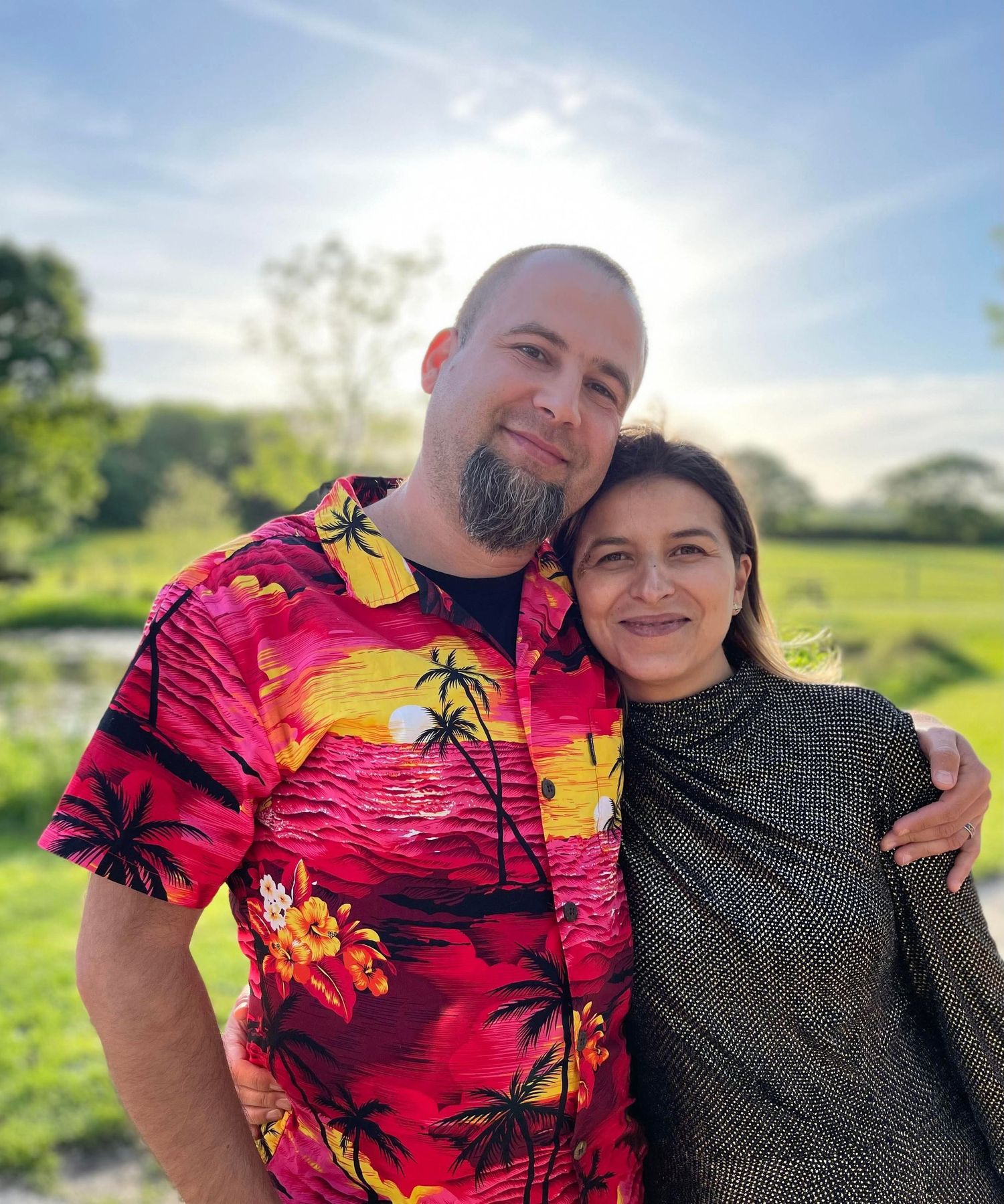 Two people pose together outdoors wearing colorful tropical print shirt and dark top.