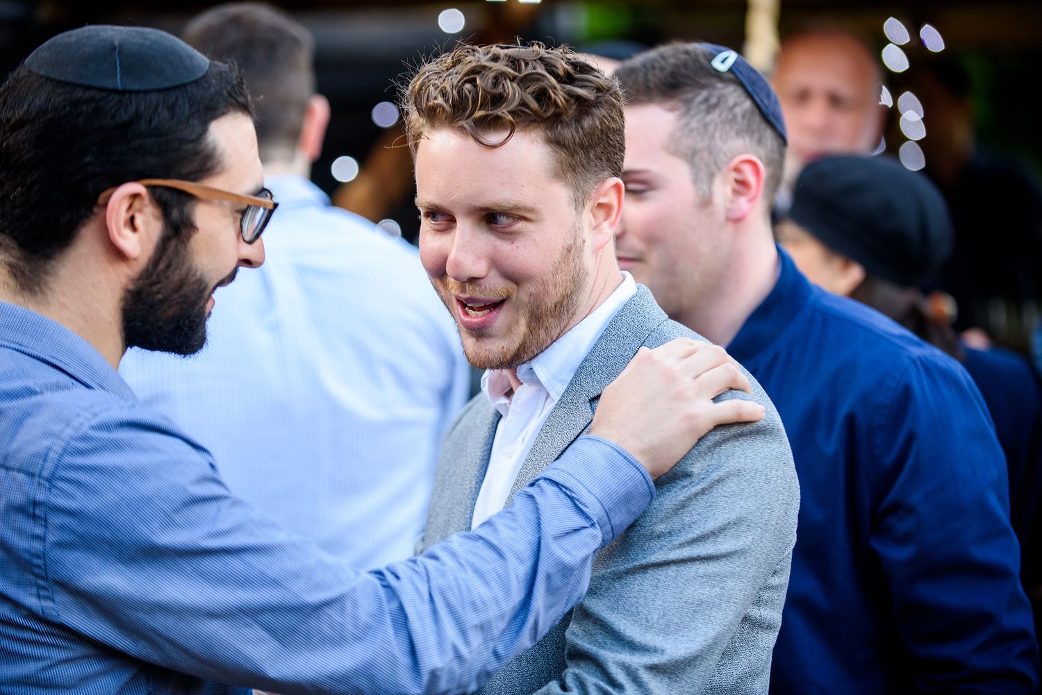 Men in suits and formal wear laugh and embrace at an outdoor gathering.