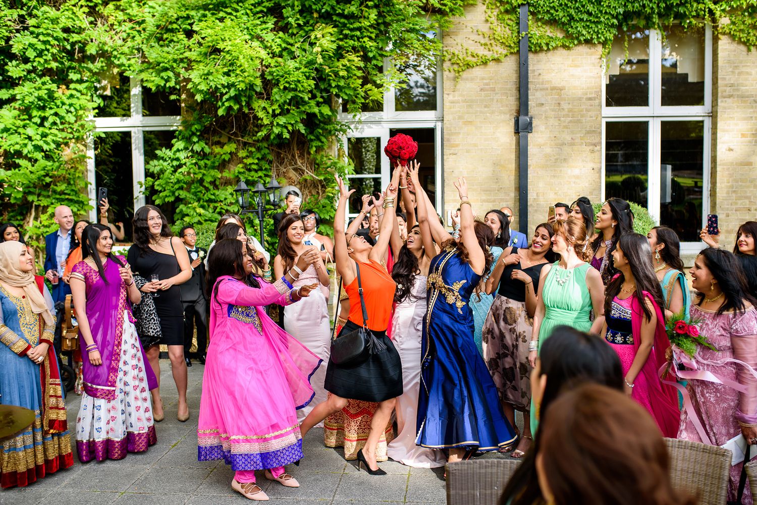 Wedding celebration crowd in bright colors gathered outside yellow building with greenery.