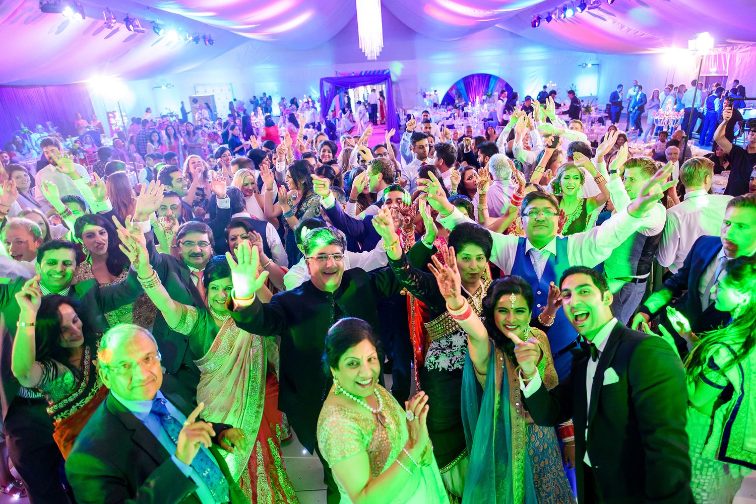 Large group of wedding guests dancing together under purple and green lighting at a festive reception.